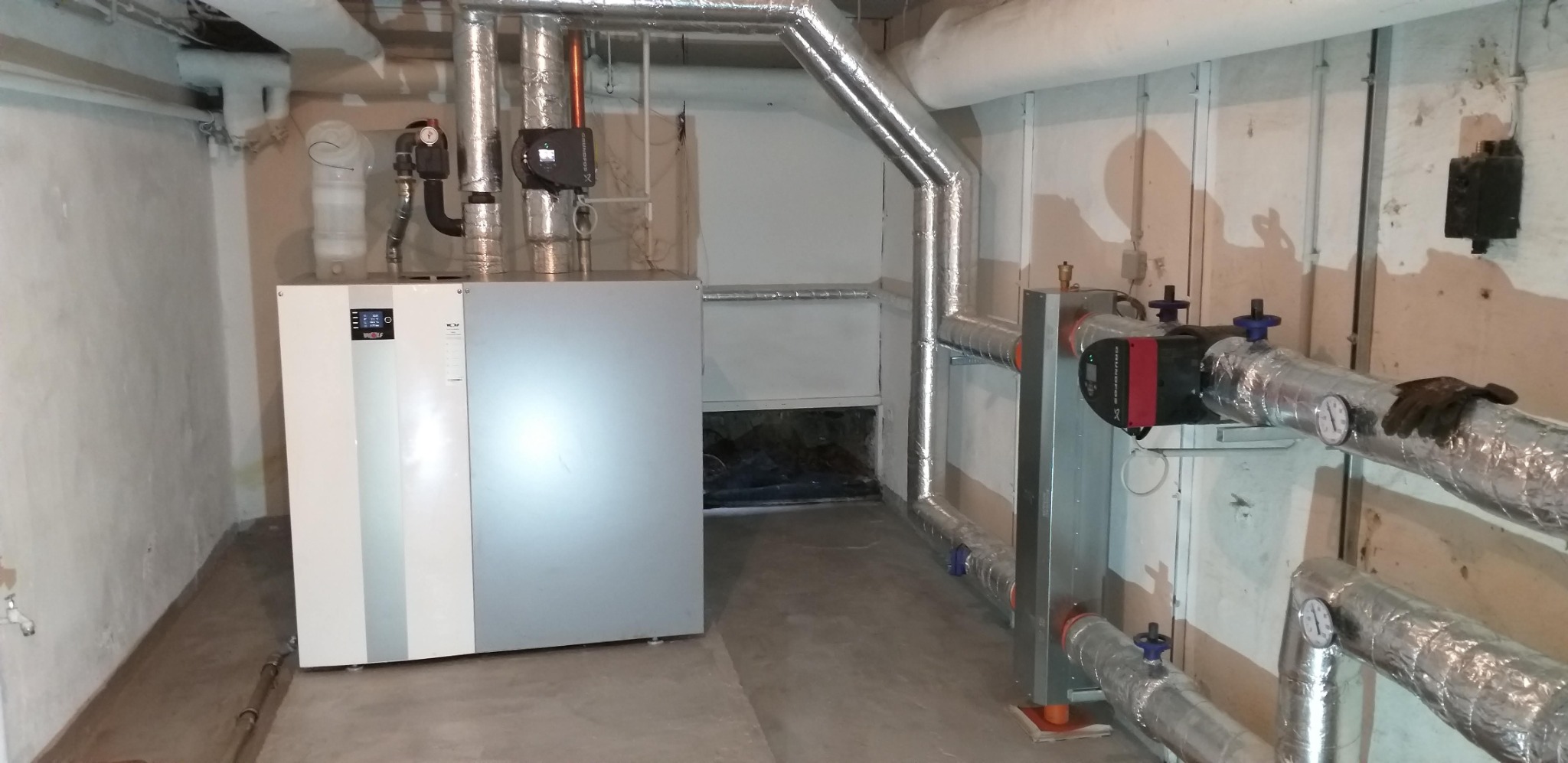 Nowoczesny piec grzewczy marki Wolf z podłączonymi, zaizolowanymi rurami, manometry oraz rękawice robocze na rurze w piwnicy.