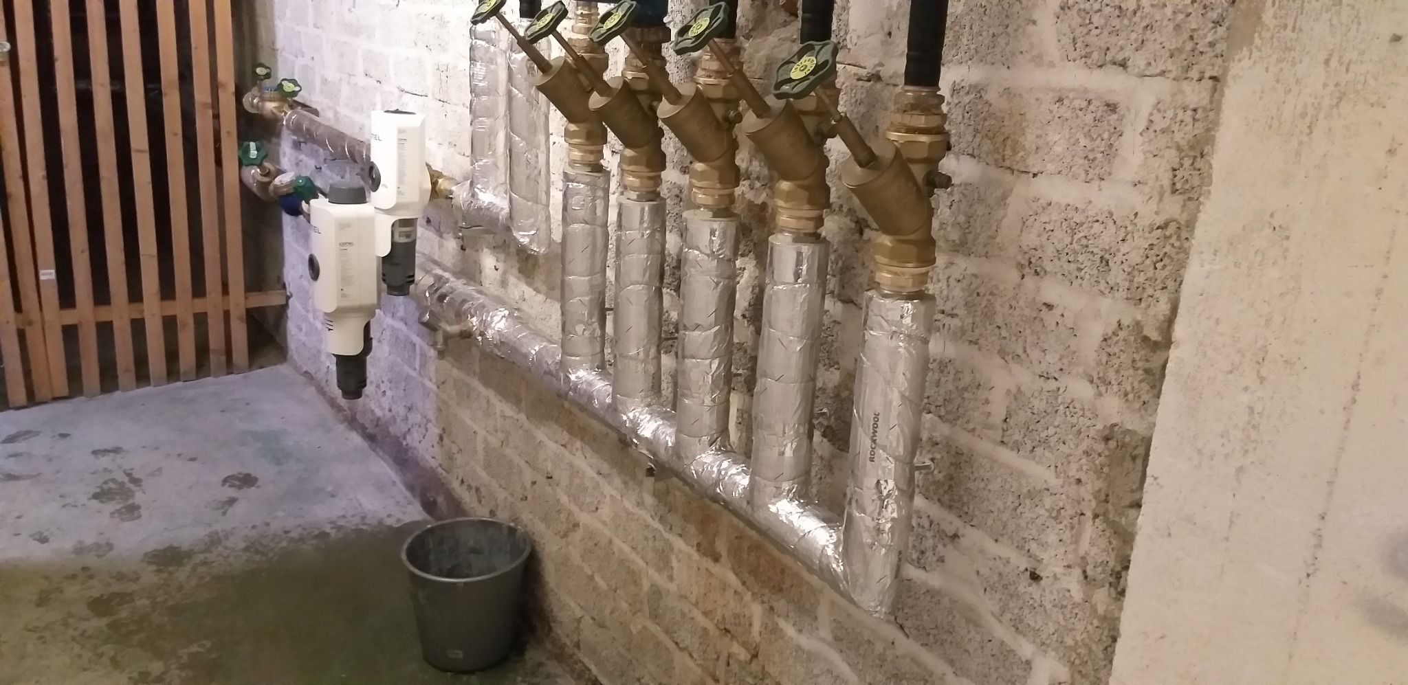 Rozdzielacz instalacji grzewczej z zaworami, rurami zaizolowanymi folią aluminiową i filtrami, zamontowany na ścianie z surowej cegły w piwnicy.