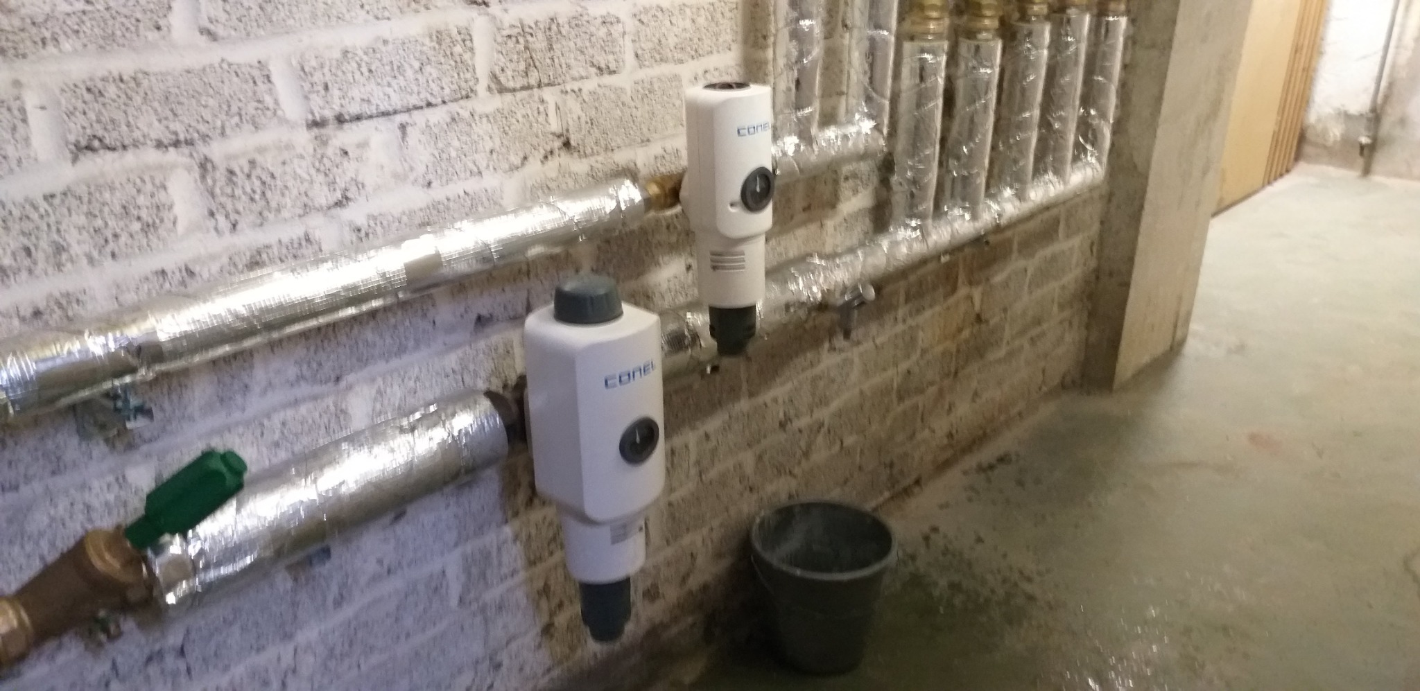 Dwa filtry magnetyczne Conel zamontowane na rurach instalacji grzewczej, z izolacją termiczną, na tle ściany z białej cegły i mokrej podłogi.