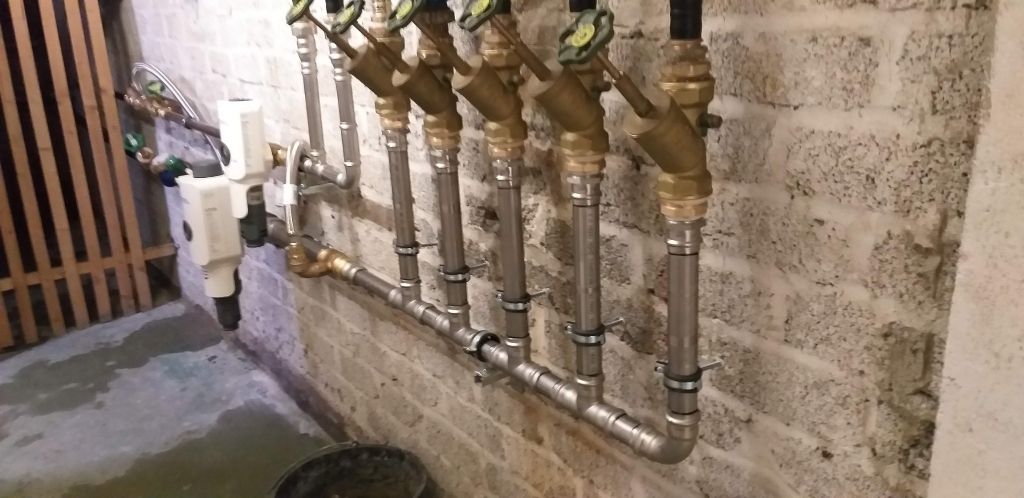 Rząd pionowych rur z zaworami, przymocowanych do ściany z bloków betonowych, z widocznymi elementami instalacji filtrującej i fragmentem drewnianej konstrukcji w tle.