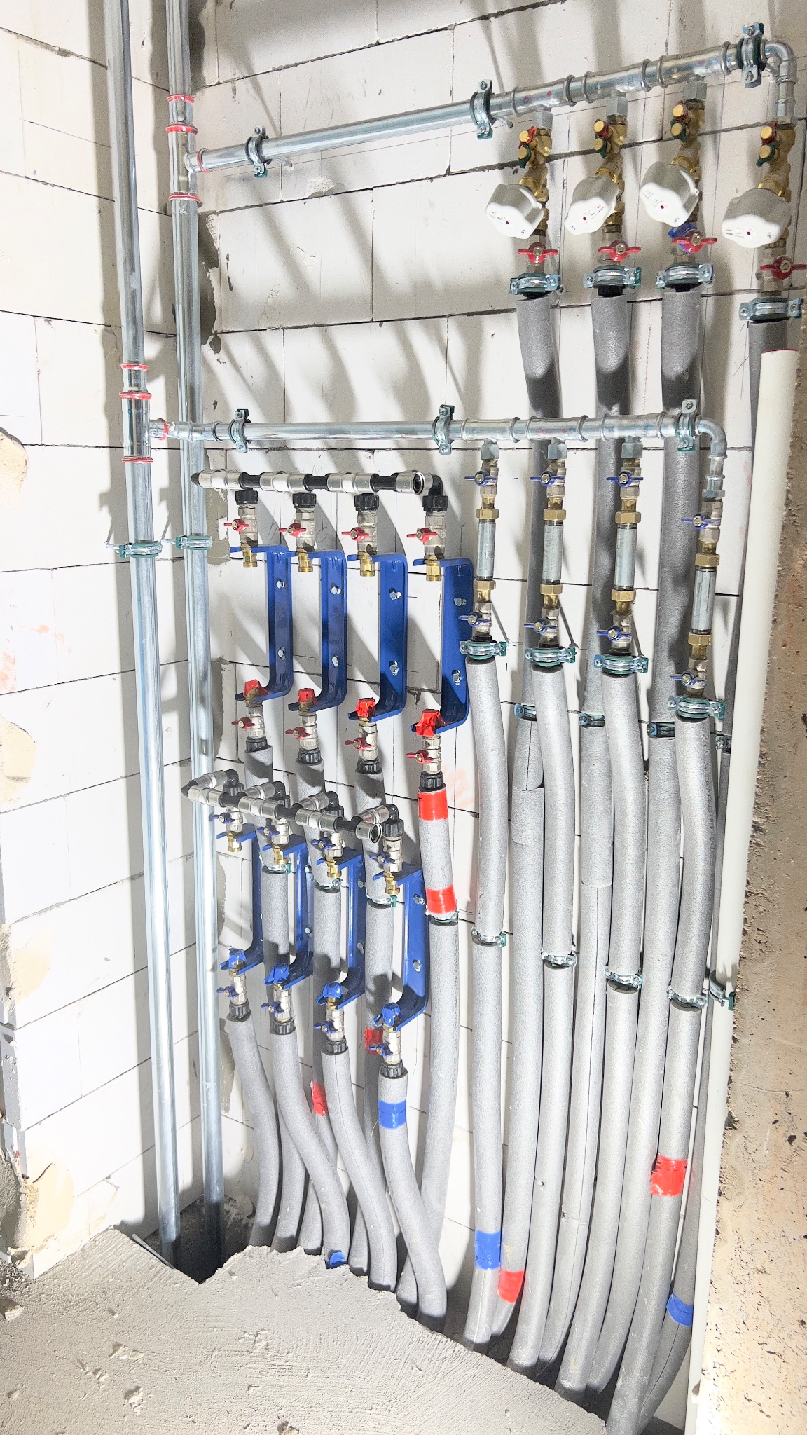 Rozdzielacz ogrzewania podłogowego z rurami w izolacji, zamontowany na ścianie z białego bloczka. Widoczne zawory, mocowania i fragment wylewki podłogowej.