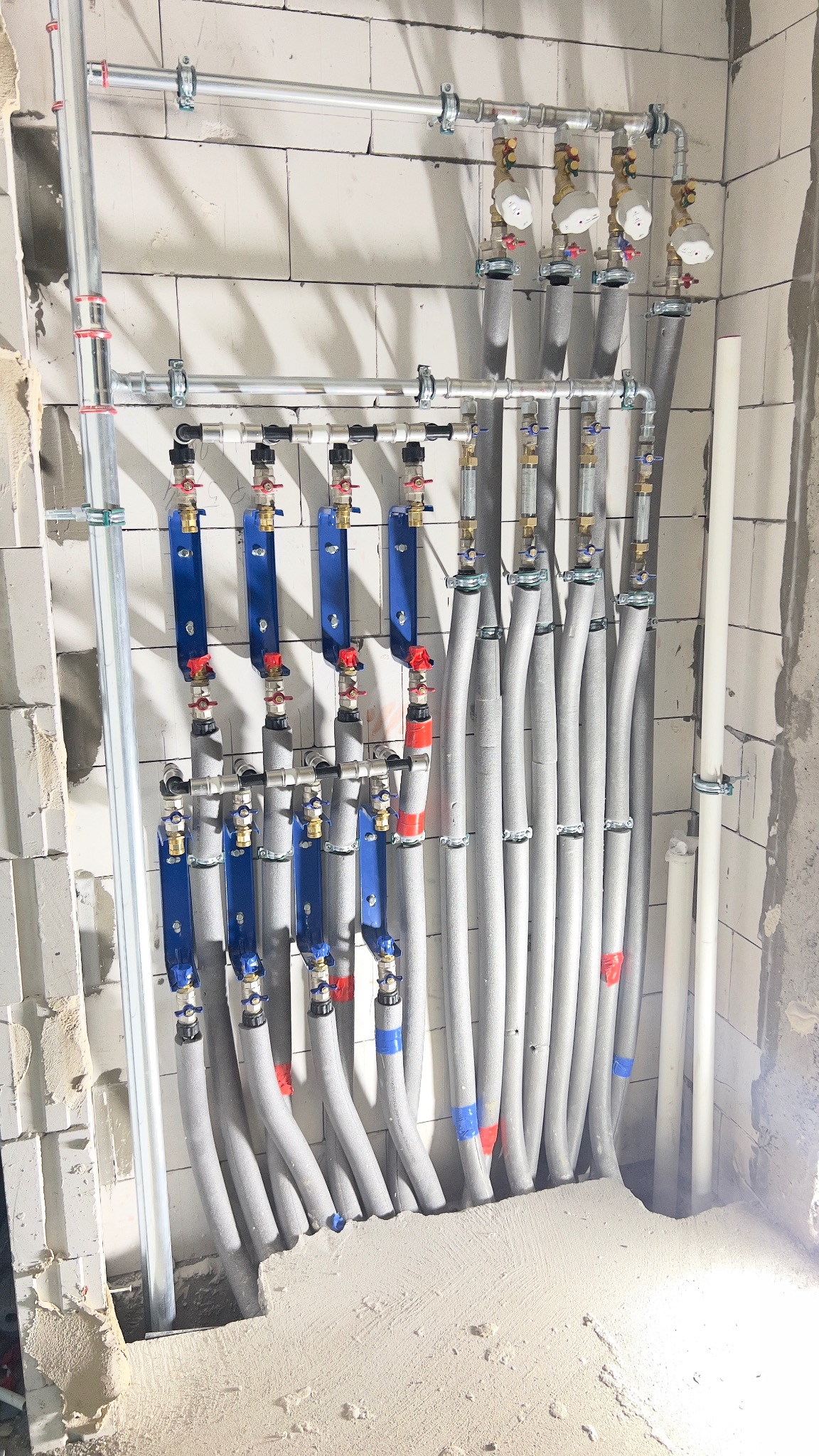 Szczegółowe ujęcie instalacji grzewczej z rurami w izolacji, zaworami i mocowaniami na surowej ścianie z betonu komórkowego. Widoczne elementy instalacji podłogowej.