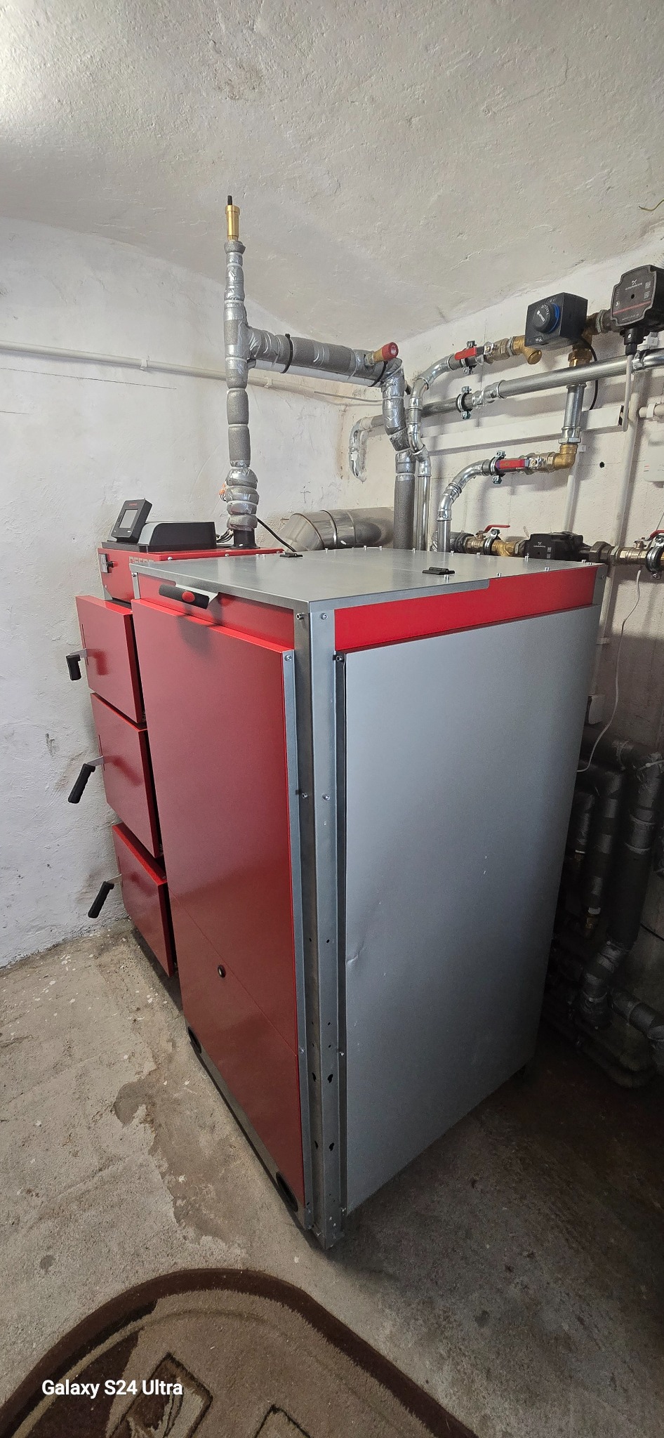 Nowoczesny piec CO z czerwonymi drzwiczkami i szarą obudową, z widoczną instalacją hydrauliczną w tle, zamontowany w kotłowni.