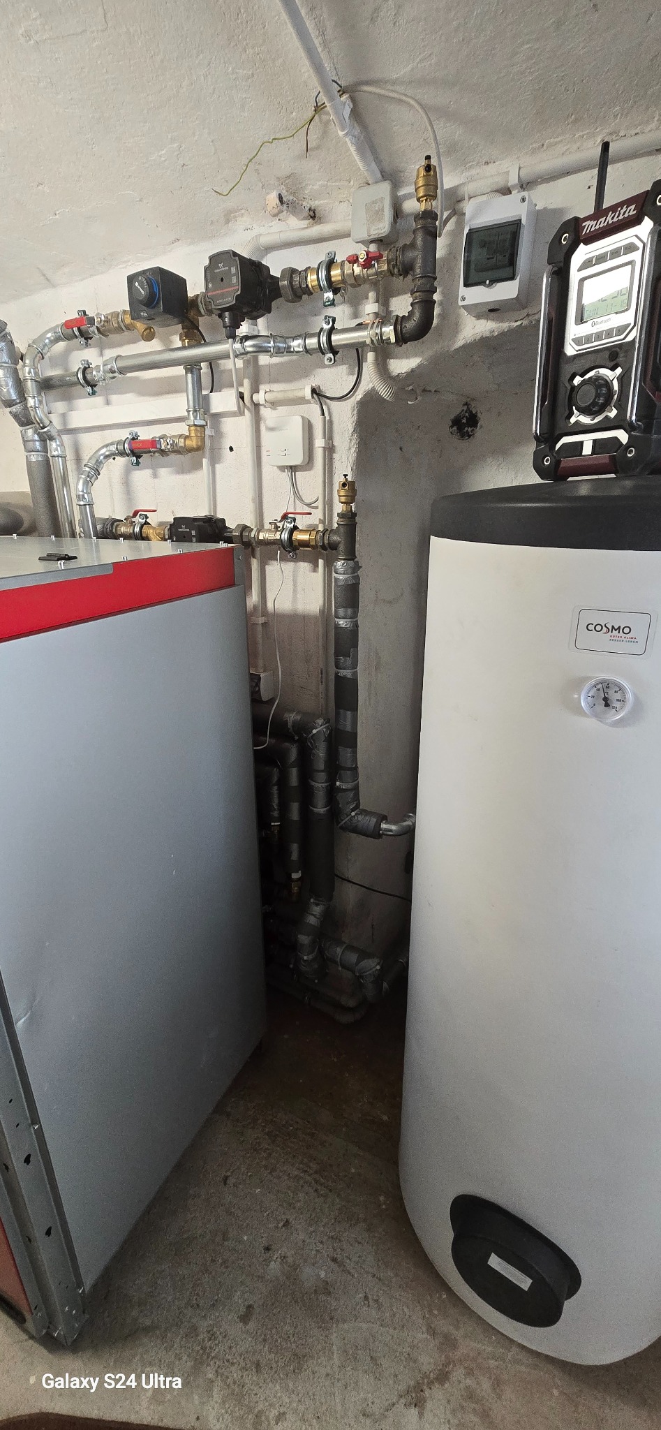 Złożona instalacja grzewcza z kotłem, zasobnikiem ciepłej wody i licznymi rurami. Widoczne elementy sterujące i izolacja termiczna na rurach.