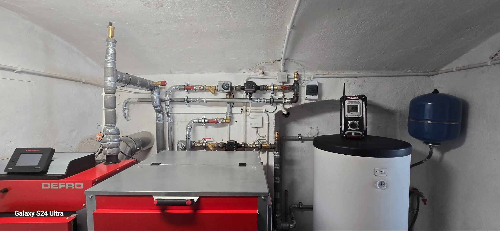 Nowoczesny piec CO Defro z instalacją, zbiornikiem i osprzętem. Widoczne rury, zawory, pompy obiegowe i miernik temperatury Makita. Całość w kotłowni o surowych ścianach.