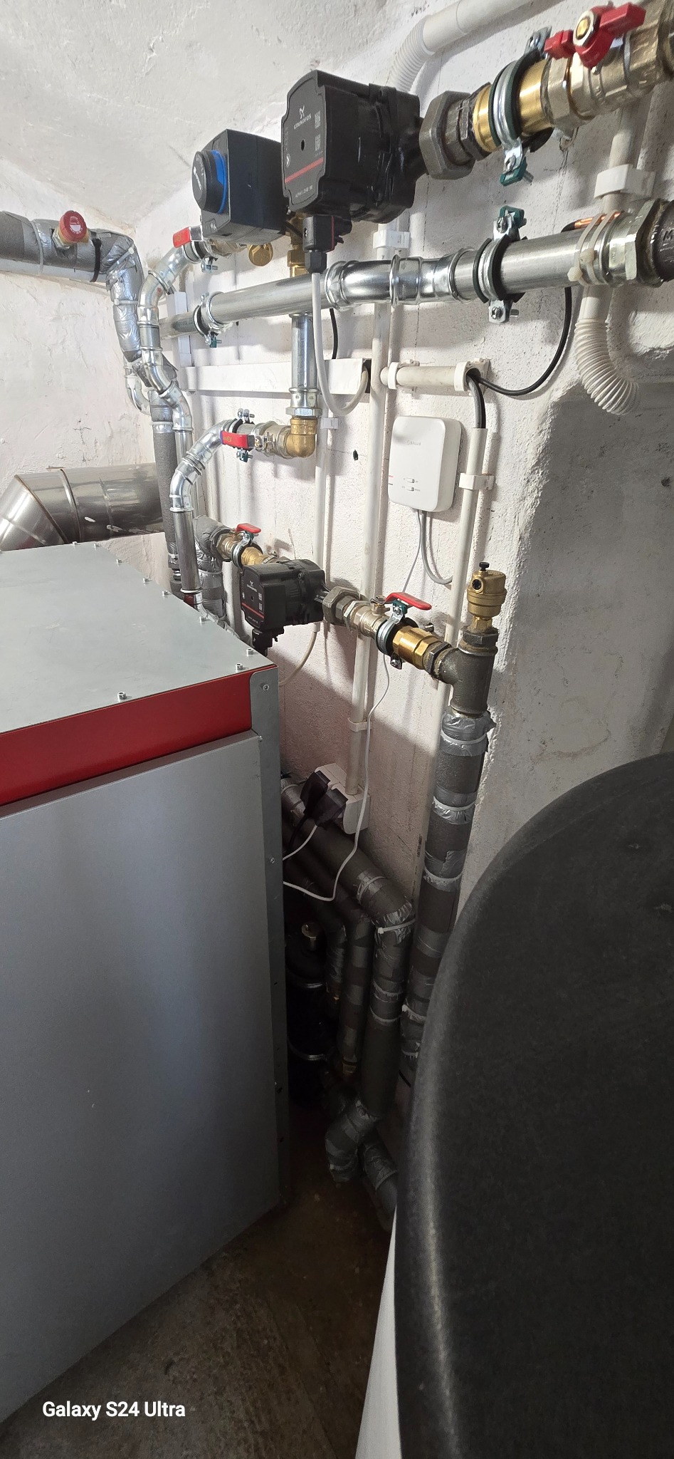 Złożona instalacja grzewcza z piecem i pompami obiegowymi, rury z zaworami, biała ściana z przewodami elektrycznymi. Widok na kotłownię z bliska.