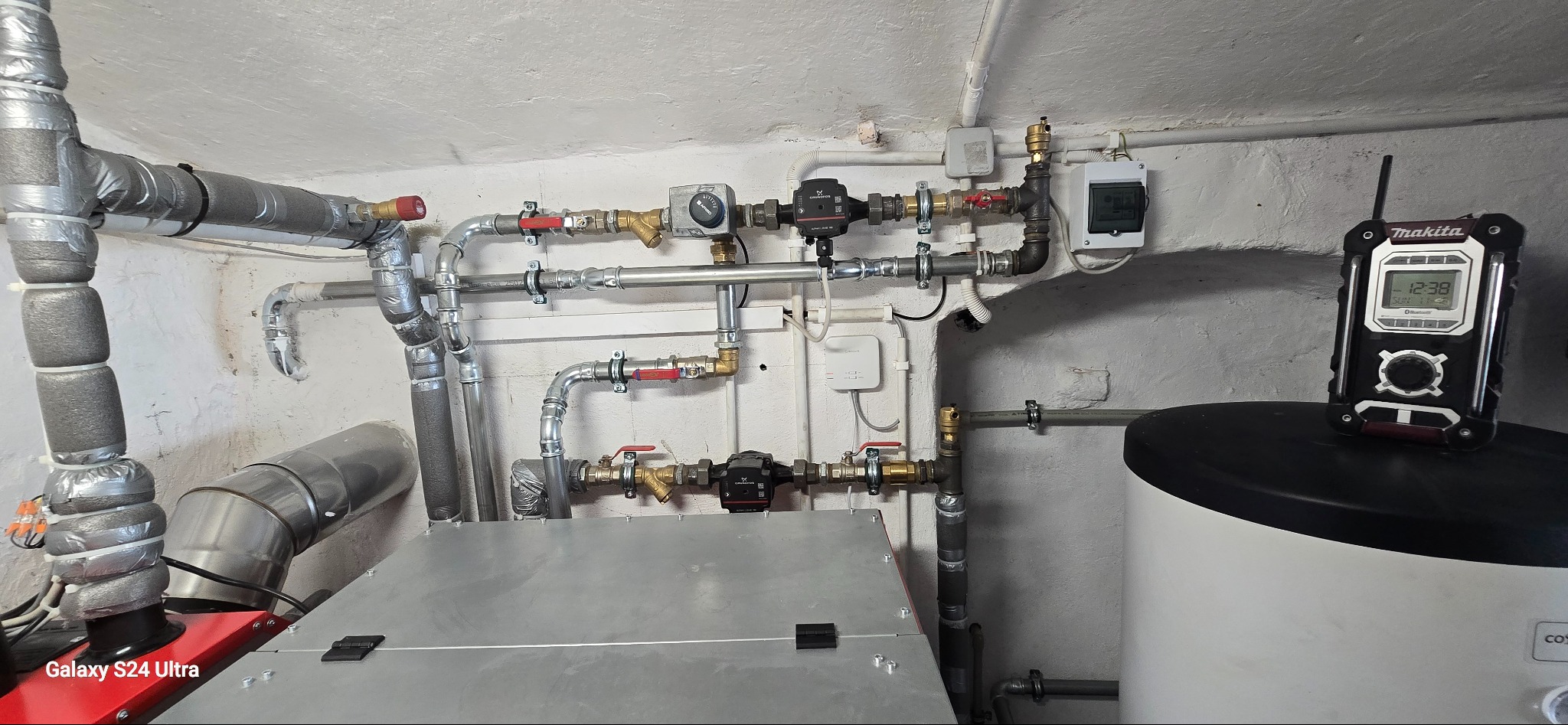 Złożona instalacja grzewcza z rurami, zaworami i pompami obiegowymi w piwnicy. Na zbiorniku CO stoi radio budowlane Makita. Widoczne elementy instalacji i osprzęt.