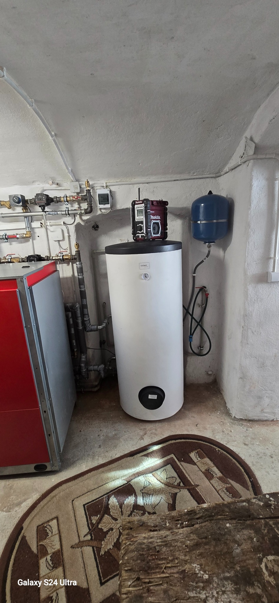 Biały bojler Cosmo z podłączonymi rurami i niebieskim zbiornikiem wyrównawczym, na górze radio Makita. Instalacja w piwnicy z dywanem na podłodze.