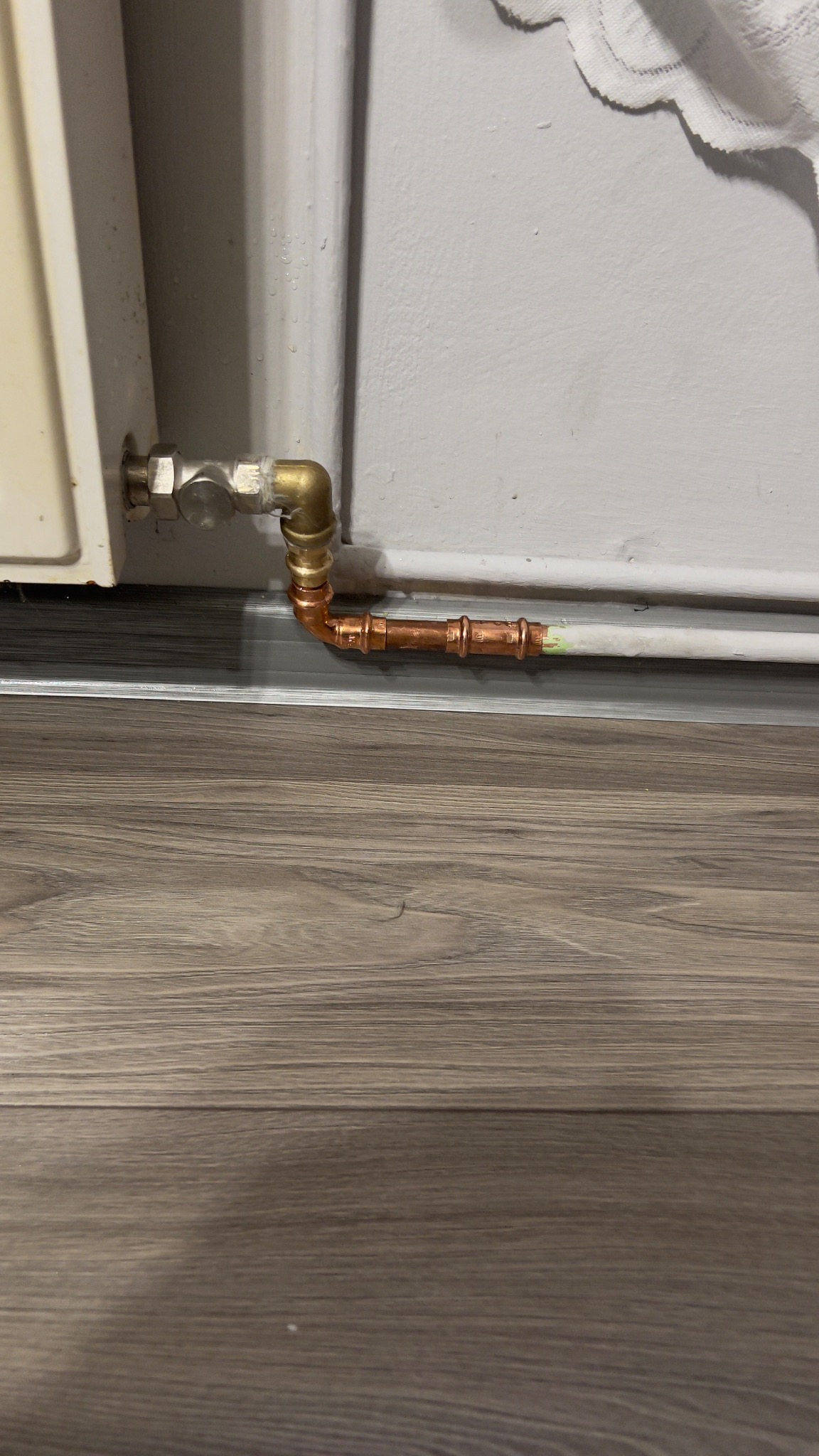 Miedziane rury połączone z grzejnikiem, widoczne złącza i fragment instalacji na tle ściany i podłogi z paneli. Detal instalacji grzewczej po montażu.
