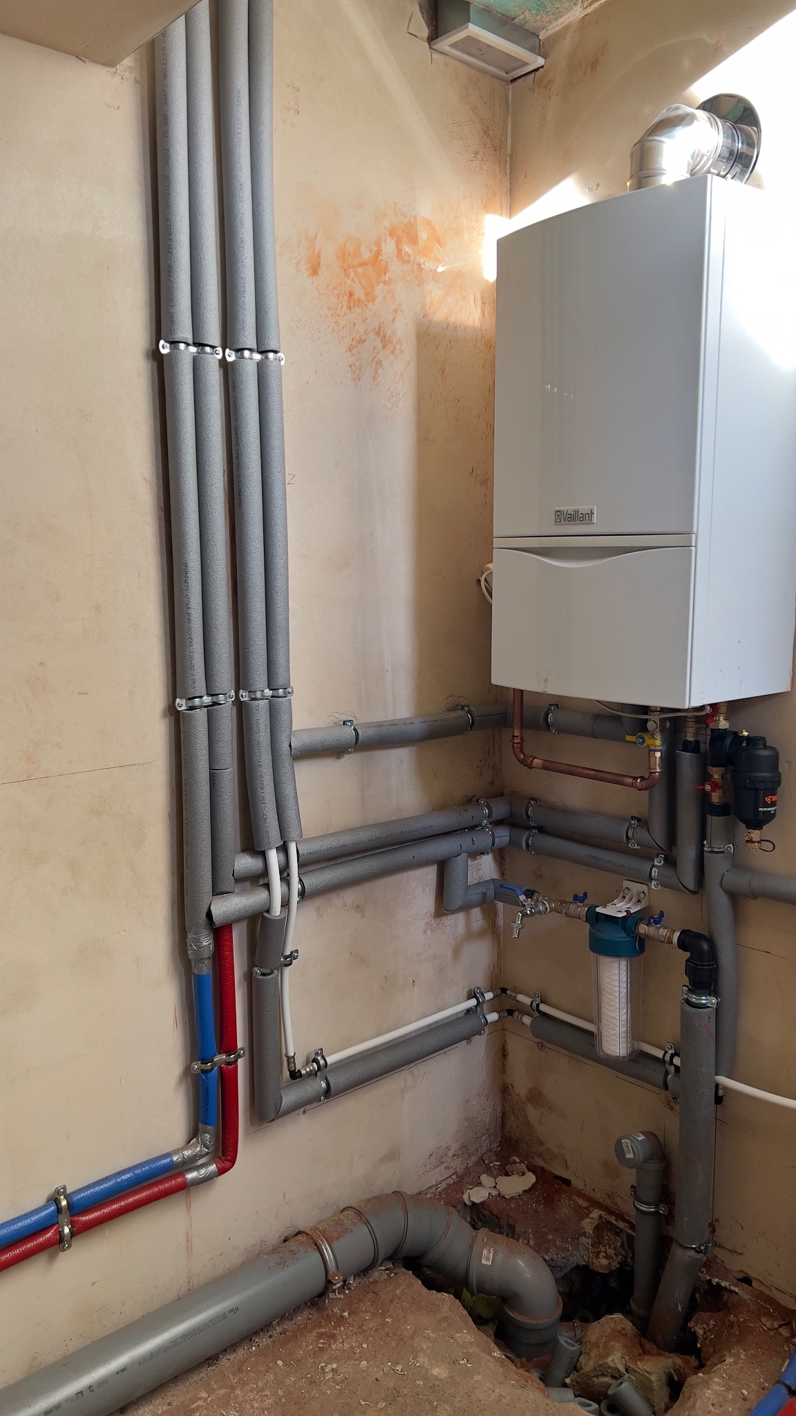 Biały piec gazowy Vaillant z podłączeniami rur miedzianych i szarych, izolowanych, na tle surowej ściany w trakcie remontu. Widoczne filtry i zawory.