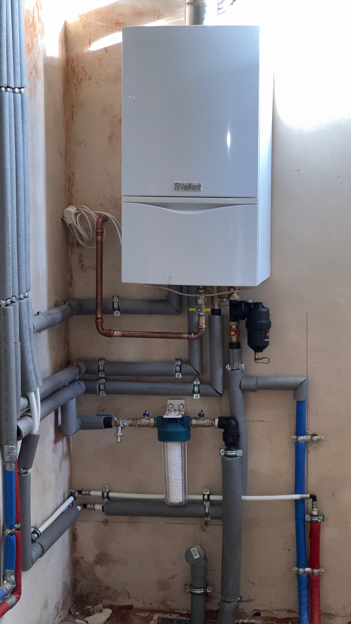 Biały piec gazowy Vaillant zamontowany na ścianie z widocznymi rurami miedzianymi i szarymi, izolowanymi przewodami. Filtr wody na pierwszym planie. Ściana w surowym stanie.