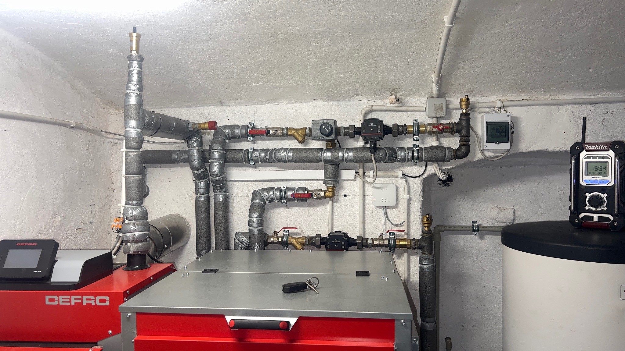 Złożona instalacja grzewcza z kotłem Defro, rurami z izolacją i zaworami, uzupełniona o radio Makita na zbiorniku, w pomieszczeniu z surowym, białym tynkiem.