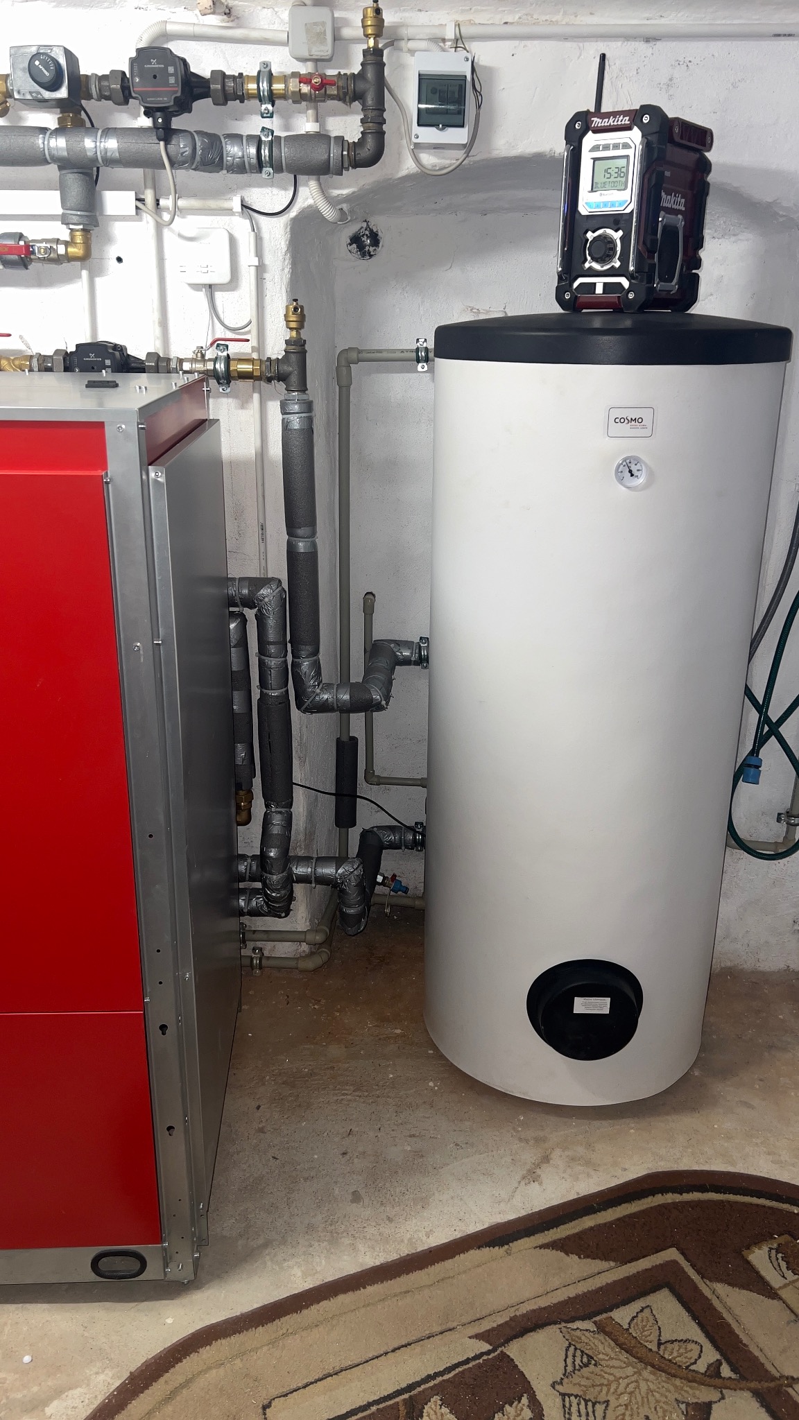 Zainstalowany piec CO z czerwonym panelem i biały zasobnik ciepłej wody Cosmo, połączone rurami. Na zasobniku radio budowlane Makita. Instalacja w piwnicy.