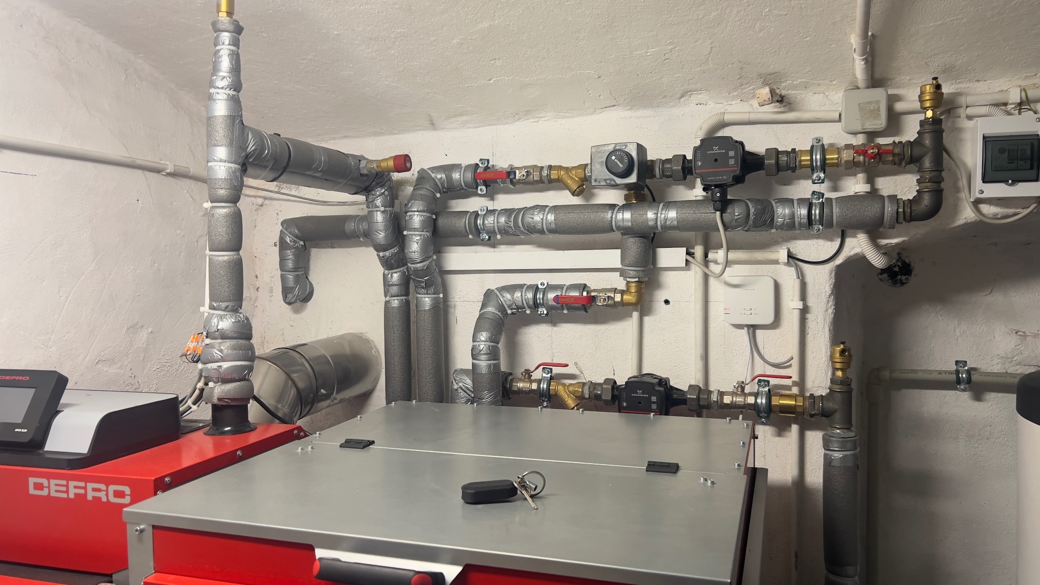 Złożona instalacja grzewcza z kotłem Defro, rurami w izolacji, zaworami i pompami obiegowymi na tle białej ściany. Klucze leżą na kotle.