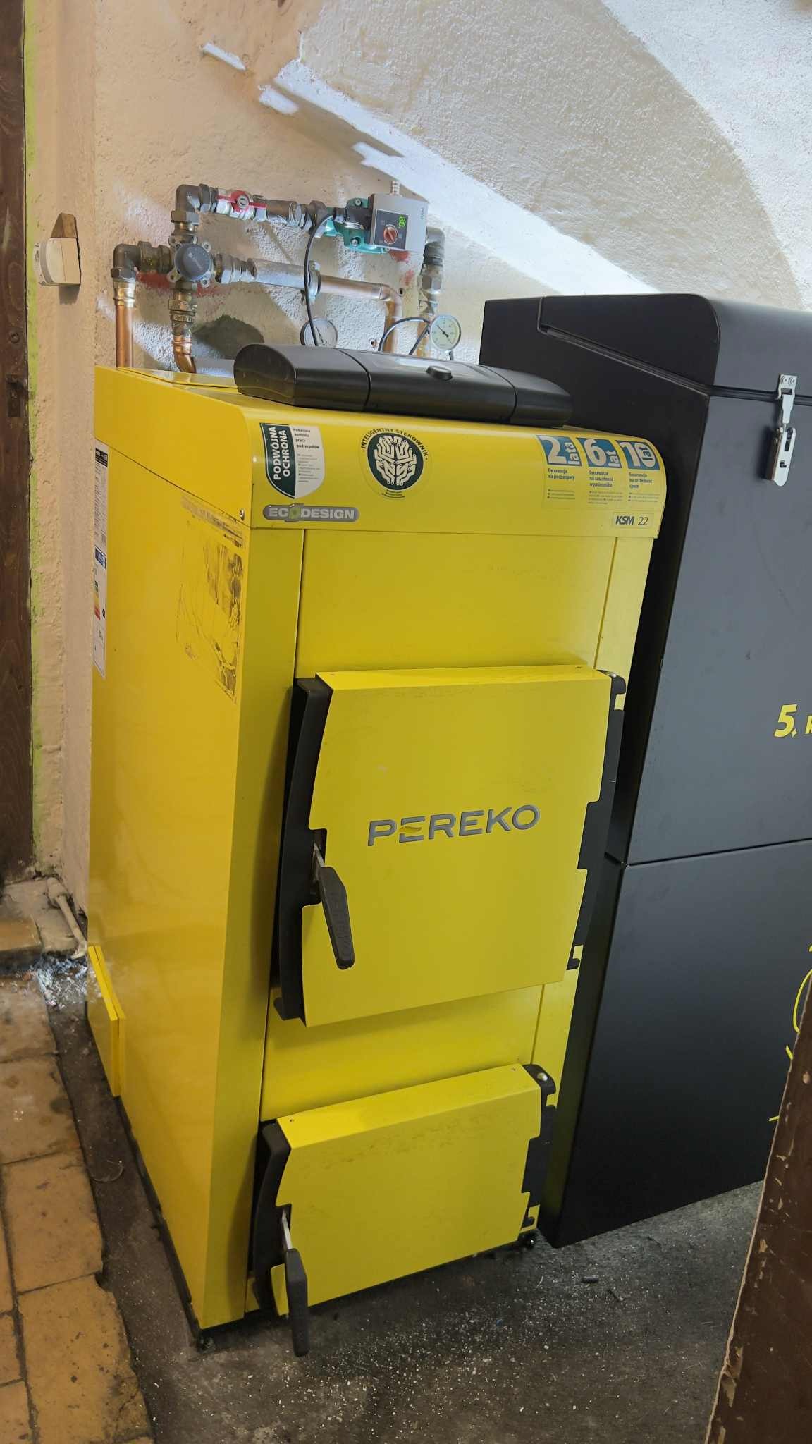 Żółty piec CO marki PEREKO z czarnymi drzwiczkami, widoczne rury instalacji grzewczej. Piec stoi w rogu pomieszczenia, obok czarny zasobnik.