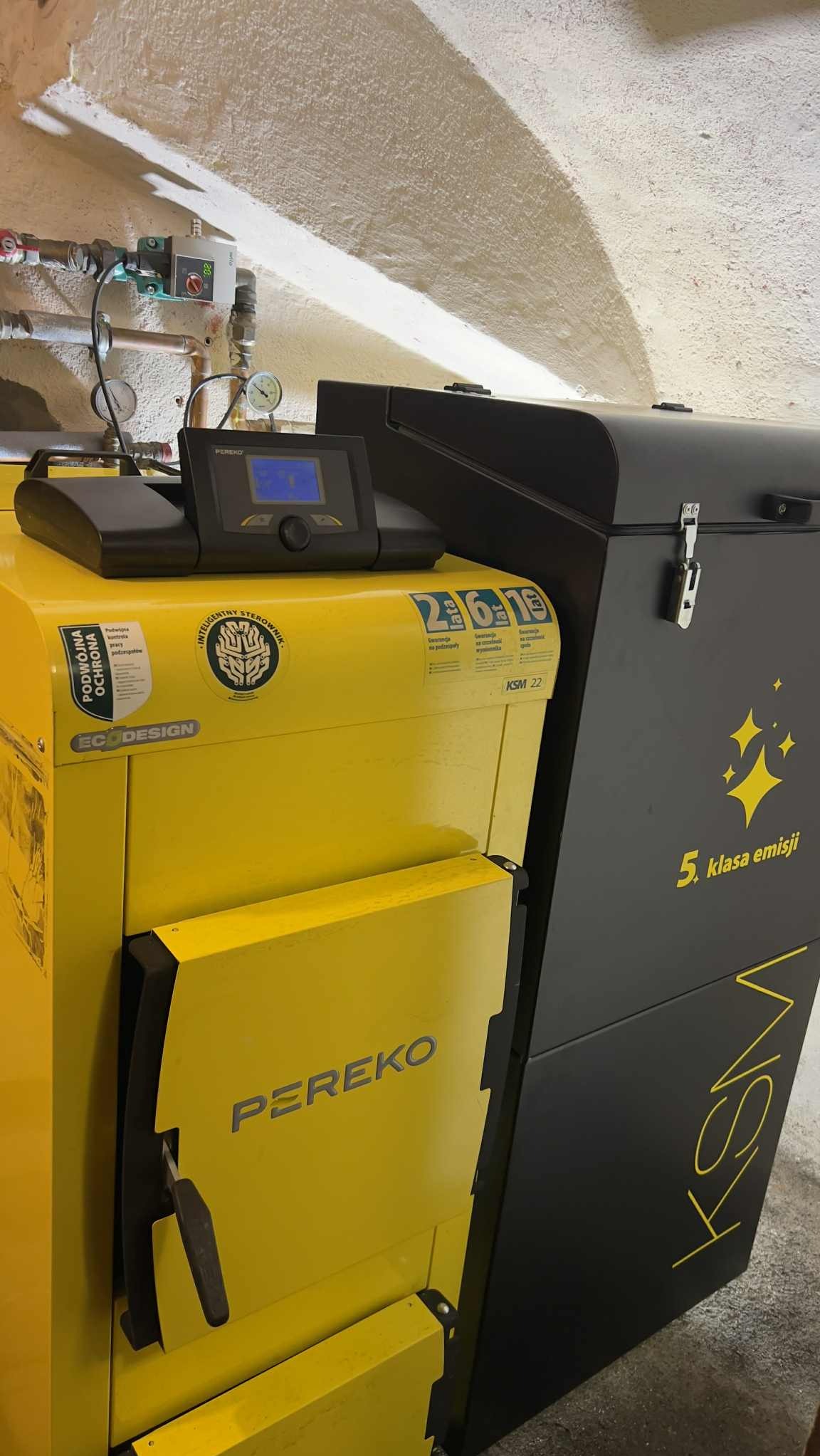 Żółto-czarny piec CO marki PEREKO KSM 22 z wyświetlaczem, naklejkami informacyjnymi i oznaczeniem klasy emisji, zainstalowany w pomieszczeniu z łukowatym sufitem.