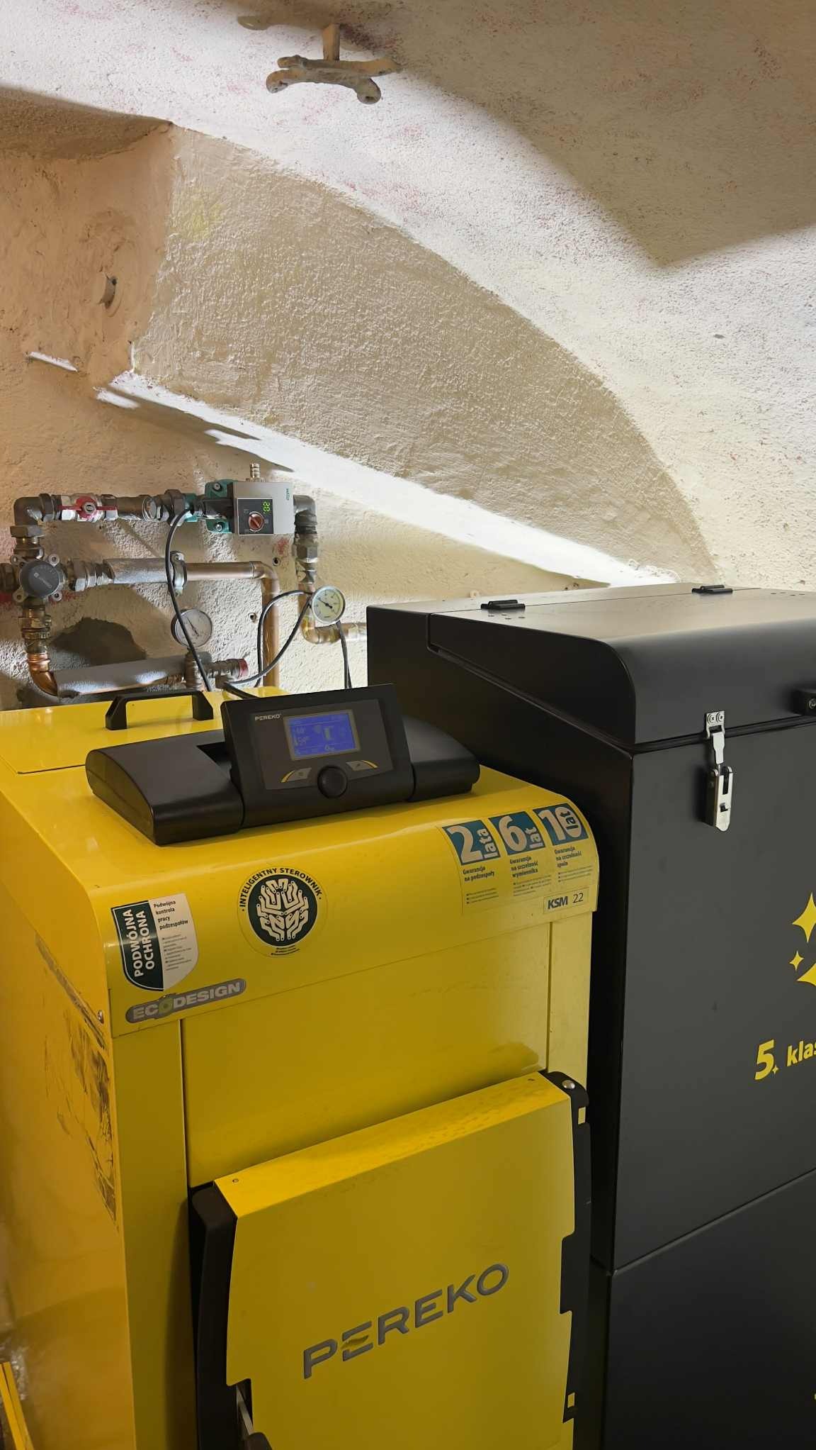 Żółty piec CO marki PEREKO z czarnym sterownikiem i zasobnikiem, widoczne rury instalacji. Ściana z fakturą baranka. Kocioł KSM 22 z klasą 5.