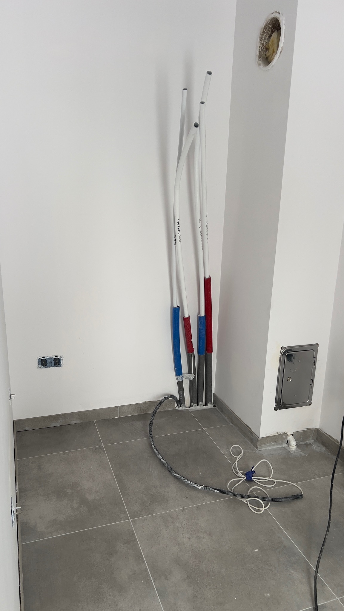 Białe rury instalacyjne z izolacją (czerwona, niebieska) wystające ze ściany w nowym wnętrzu. Widoczne gniazdka elektryczne i szara podłoga.