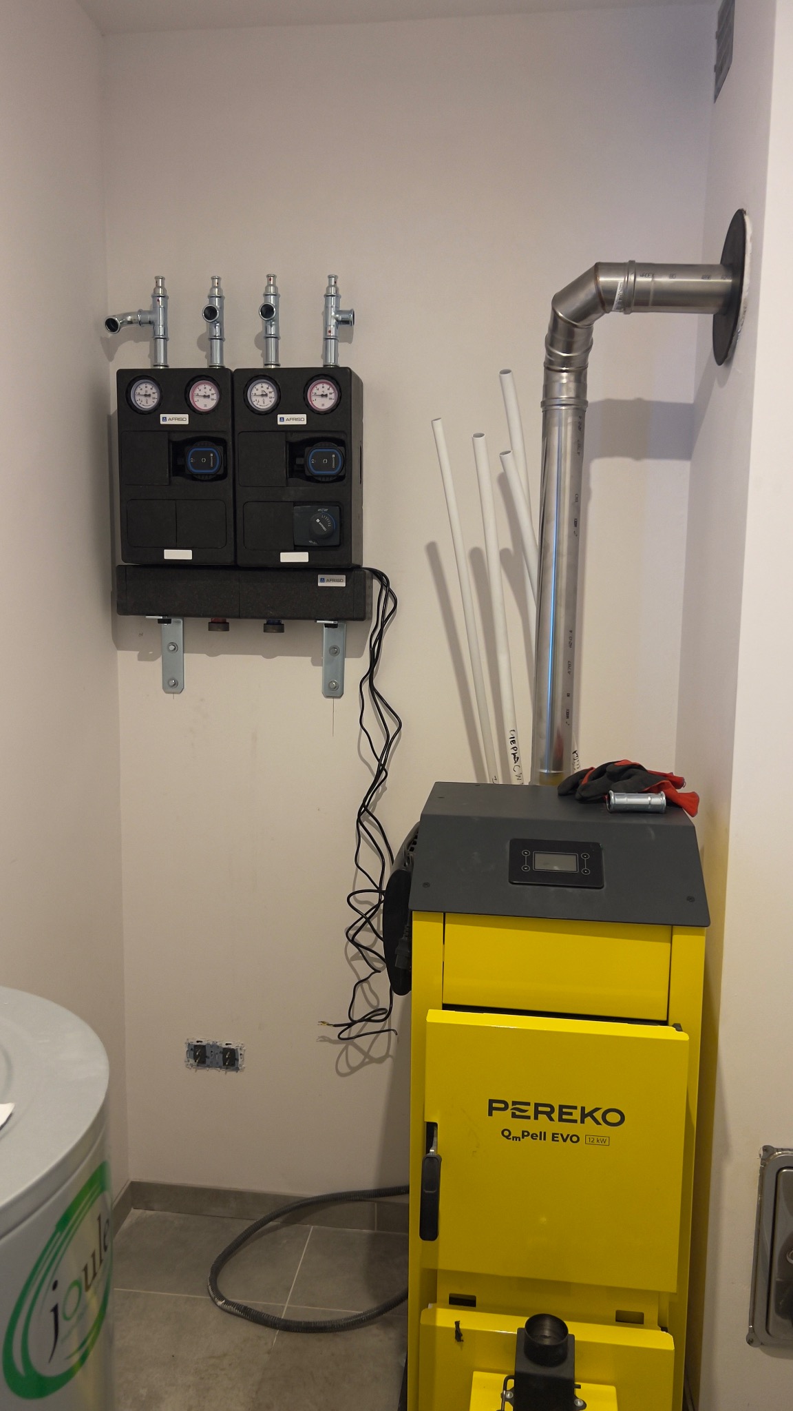 Żółty piec Pereko z podłączoną instalacją grzewczą i czarnym rozdzielaczem na ścianie. Widoczne rury, manometry i przewody. Świeżo zamontowany system grzewczy.