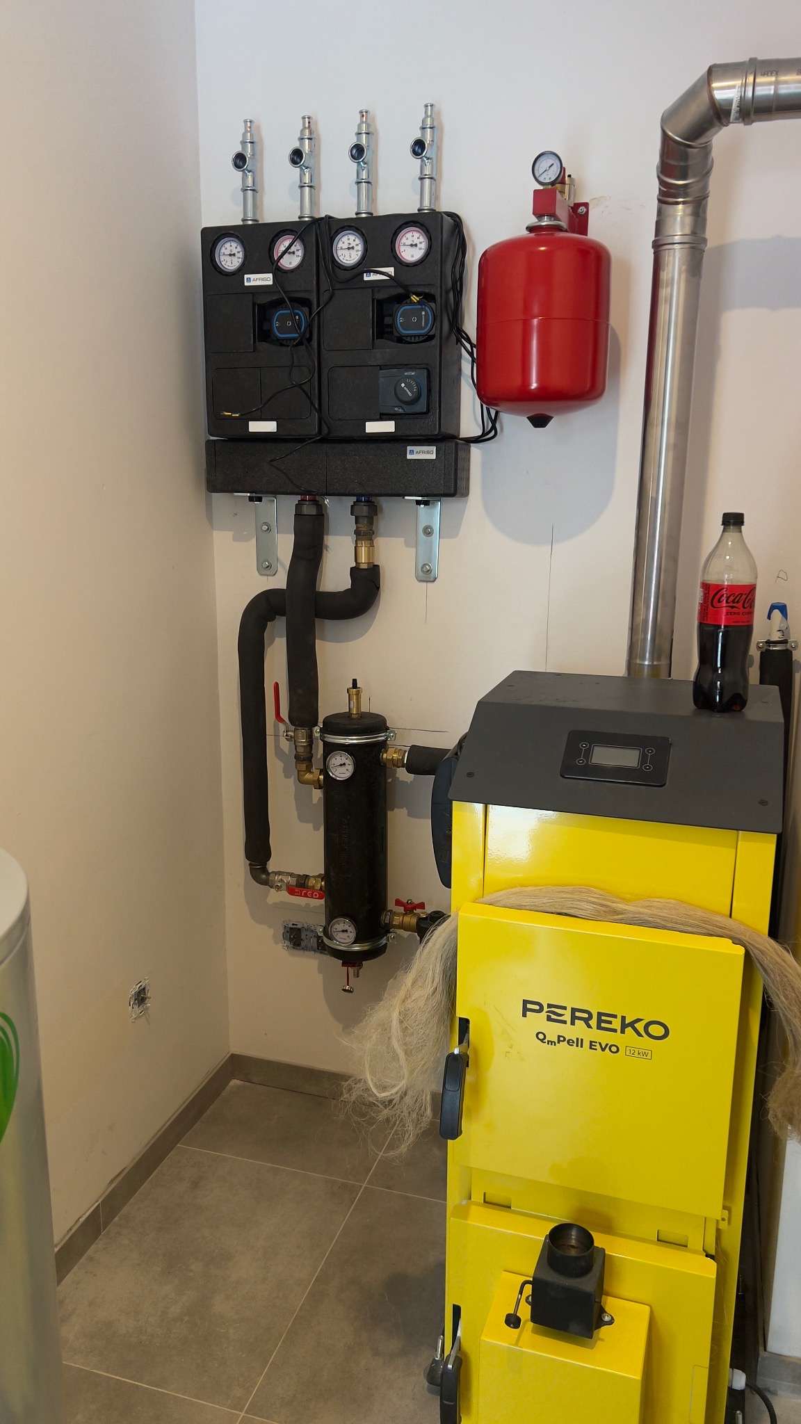 Żółty piec Pereko z instalacją CO w kotłowni w Rybnicy. Widoczne czarne rury, manometry i czerwony zbiornik wyrównawczy. Na piecu stoi butelka Coca-Coli.