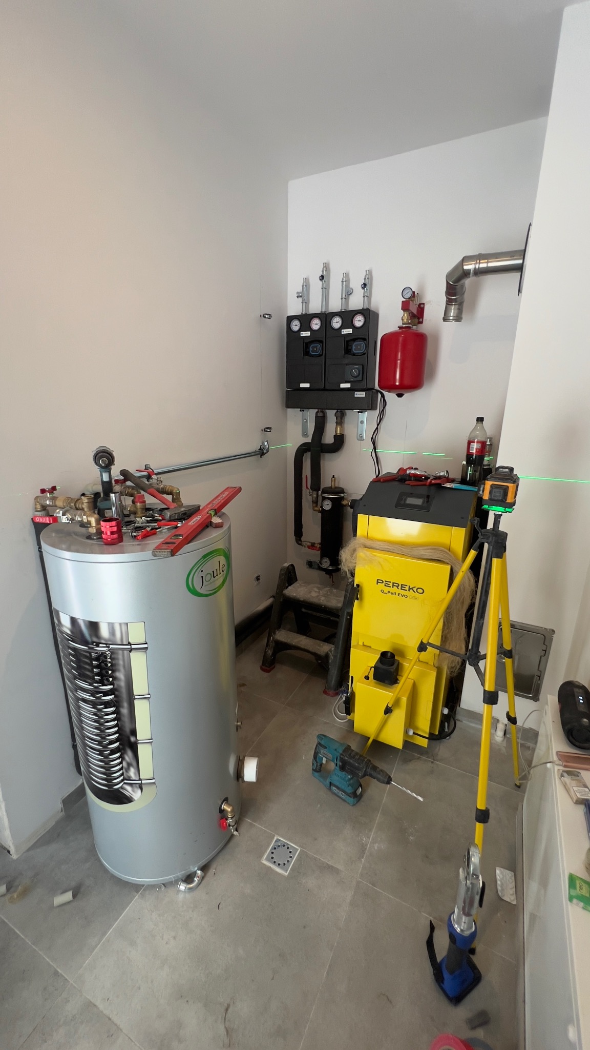Montaż kotła Pereko Alpha EVO i zasobnika Joule z narzędziami, poziomica laserowa na statywie, instalacja CO w kotłowni, szara podłoga z odpływem.