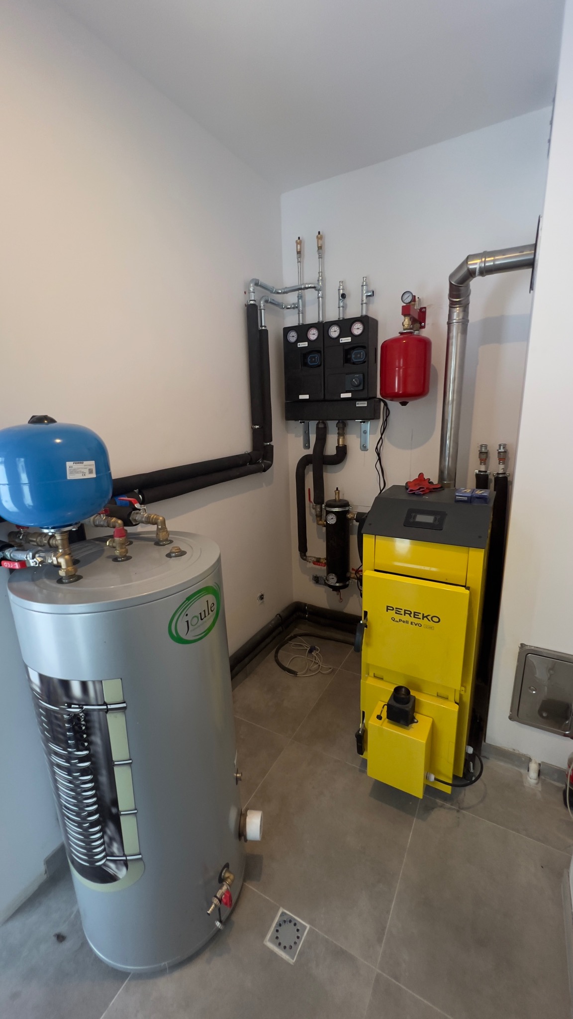 Żółty piec Pereko Q-Pell EVO i szary zasobnik Joule z niebieskim naczyniem wzbiorczym w kotłowni z szarą podłogą. Widoczne rury i czarna armatura.