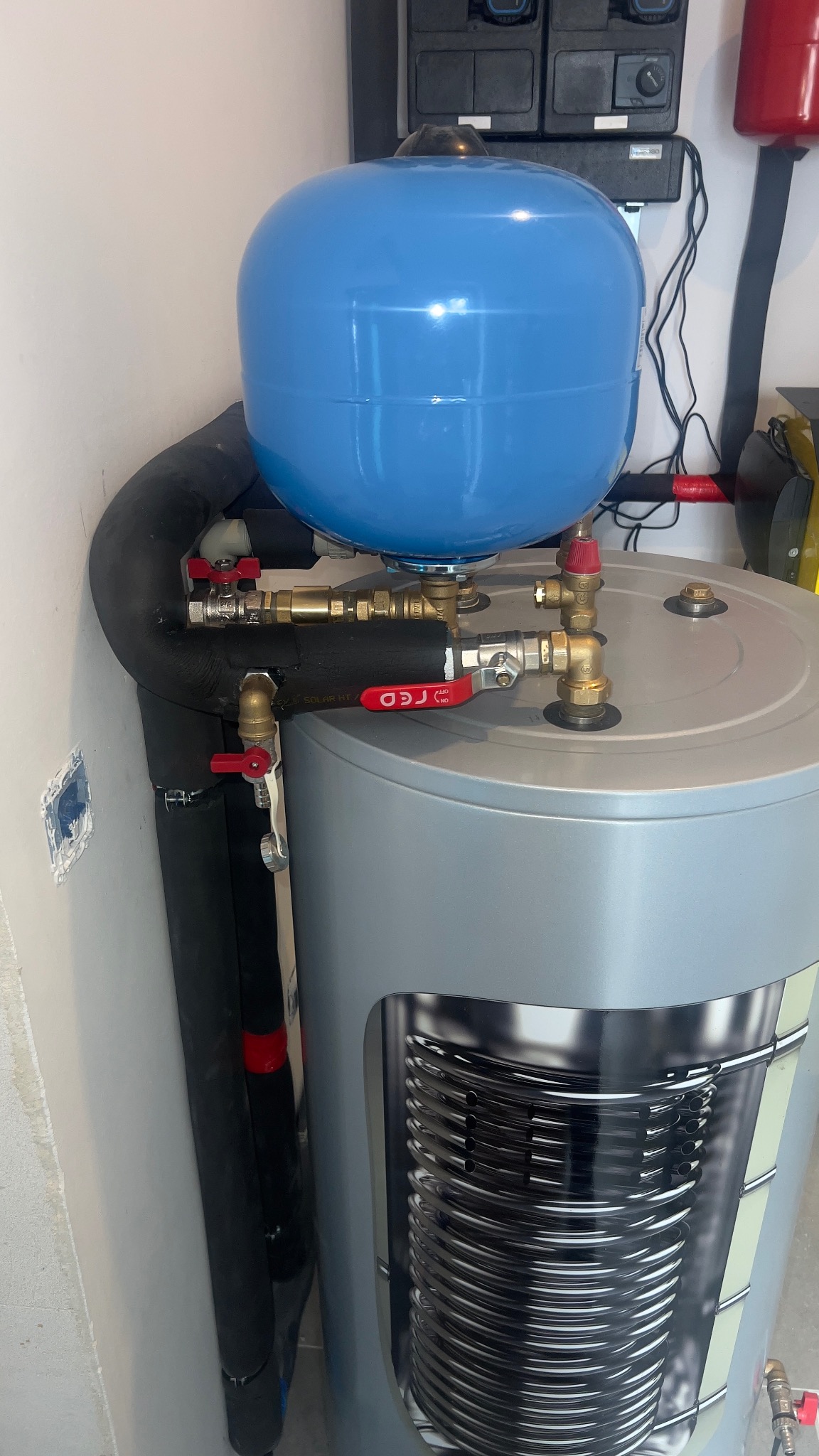 Szary bojler z niebieskim zbiornikiem wyrównawczym, widoczne rury i zawory z czerwonymi uchwytami. Elementy instalacji grzewczej w pomieszczeniu technicznym.