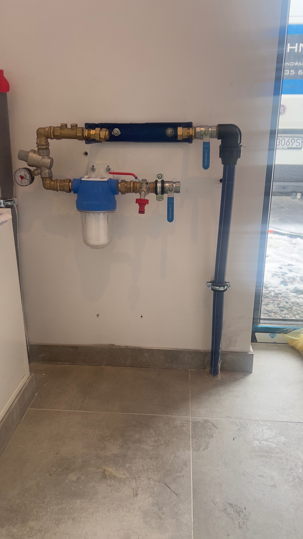 Zamontowany filtr wody z niebieską obudową i rurami miedzianymi na białej ścianie, widoczne zawory i manometr. Szare płytki podłogowe.
