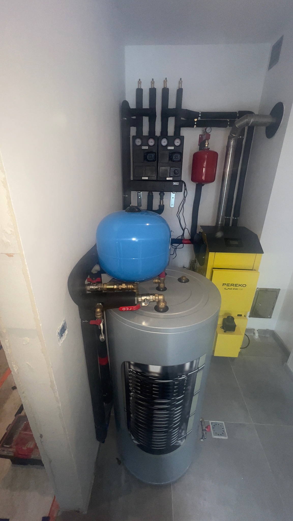 Zmontowany system grzewczy z piecem Pereko, zbiornikiem akumulacyjnym i osprzętem w kotłowni. Widoczne rury, zawory i elementy sterujące.