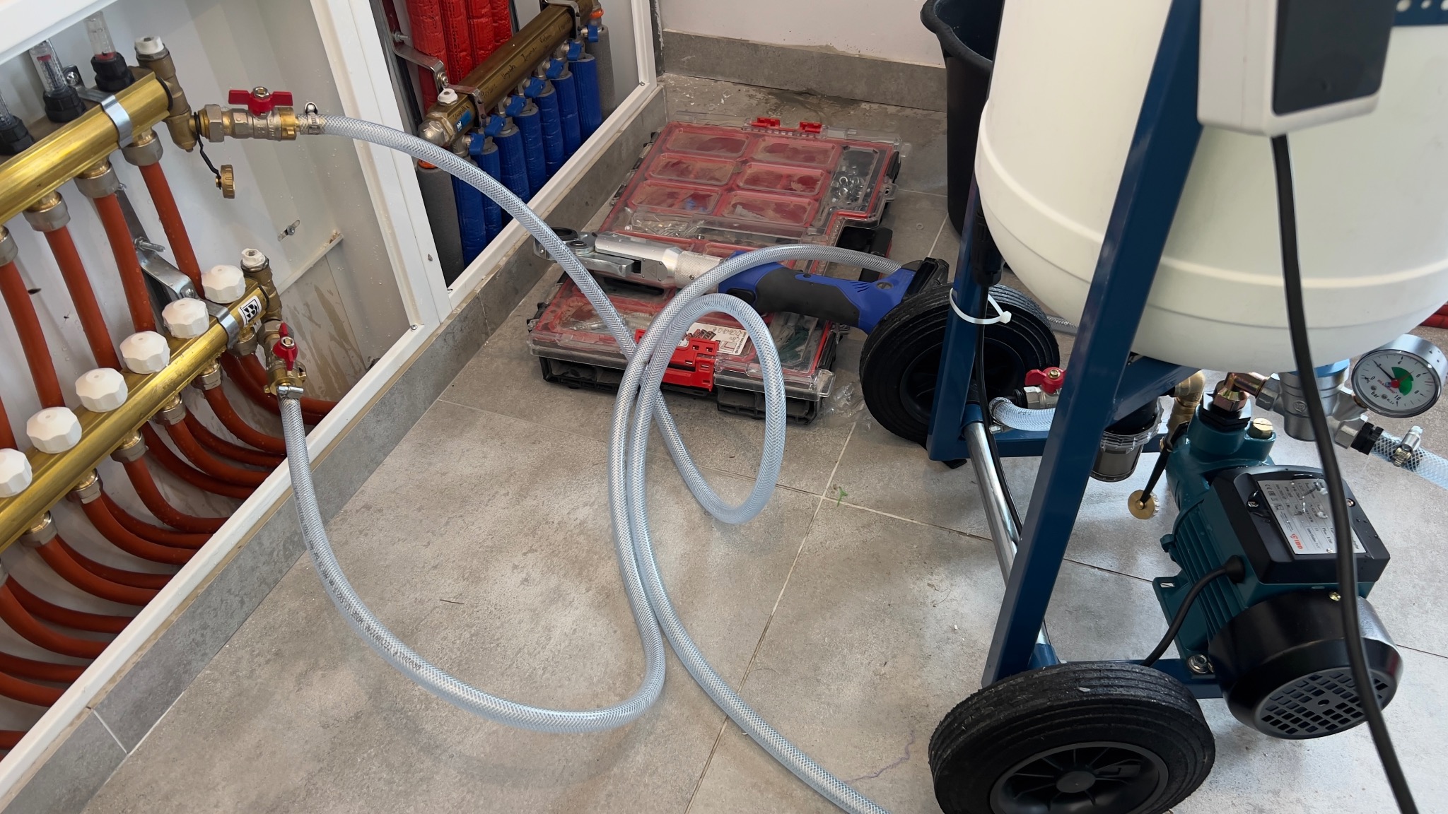Podłączenie węża do rozdzielacza ogrzewania podłogowego, widoczny manometr i narzędzia w tle. Instalacja w trakcie testów szczelności.