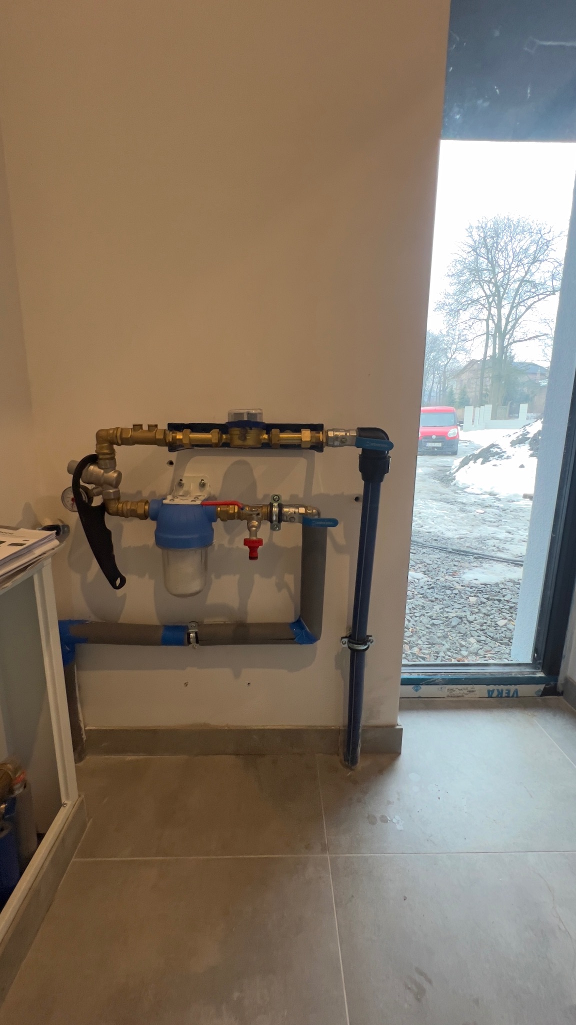 Złożona instalacja hydrauliczna z filtrem i zaworami, zamontowana na ścianie w nowym budynku. Widoczne rury miedziane i plastikowe, elementy mocujące. Za oknem widok na zimowy krajobraz.