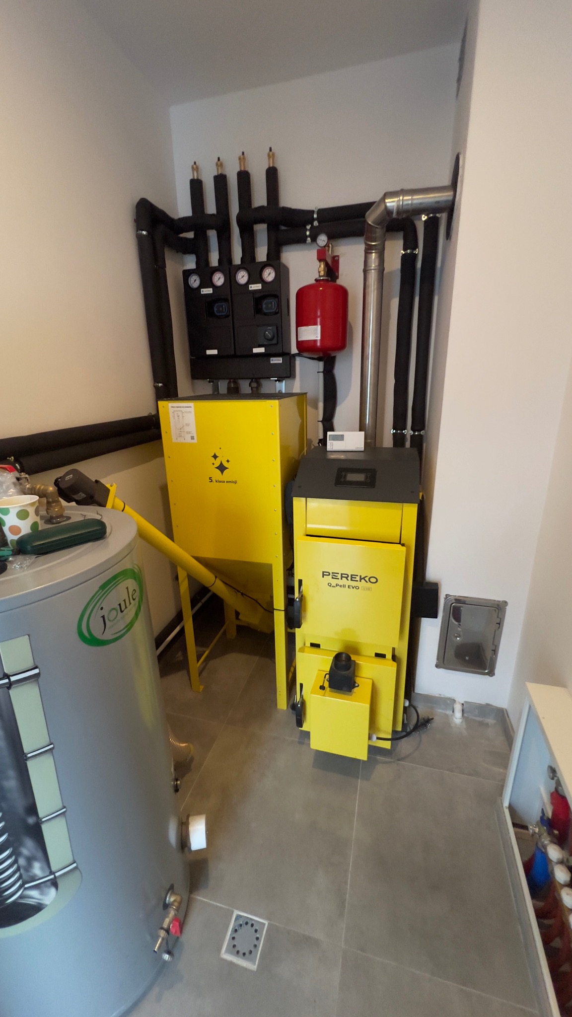 Żółty piec Pereko Q-Pell Evo z zasobnikiem, instalacja CO z czarnymi rurami, szary bojler Joule w kotłowni z szarą podłogą z płytek. Widok na kompletną instalację grzewczą.