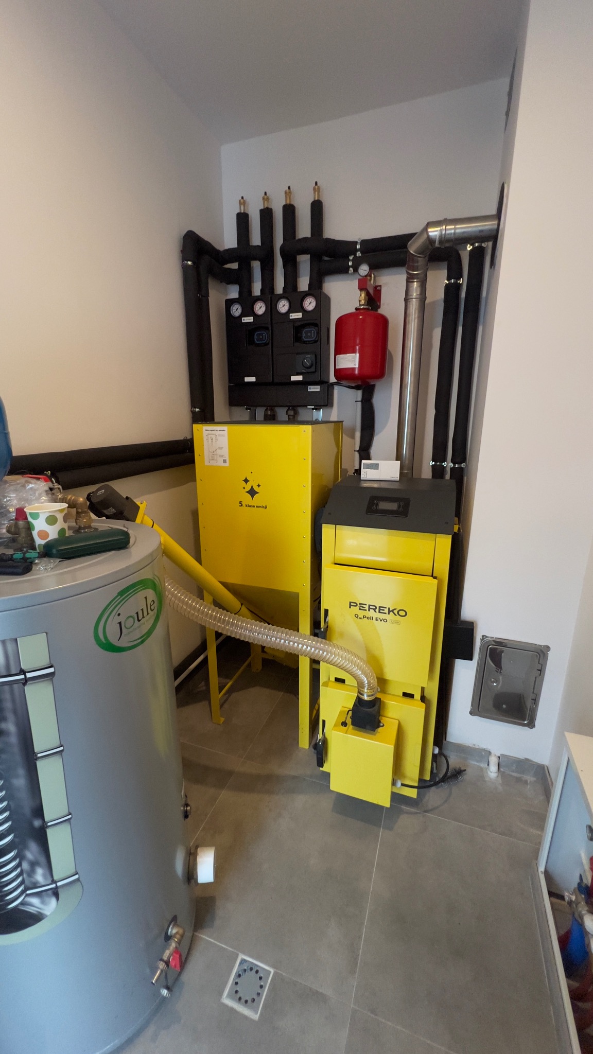 Żółty piec Pereko Q.Pell EVO 12kW z podajnikiem, zbiornik Joule i czarne rury instalacji CO w kotłowni na szarych płytkach. Widok z dołu.