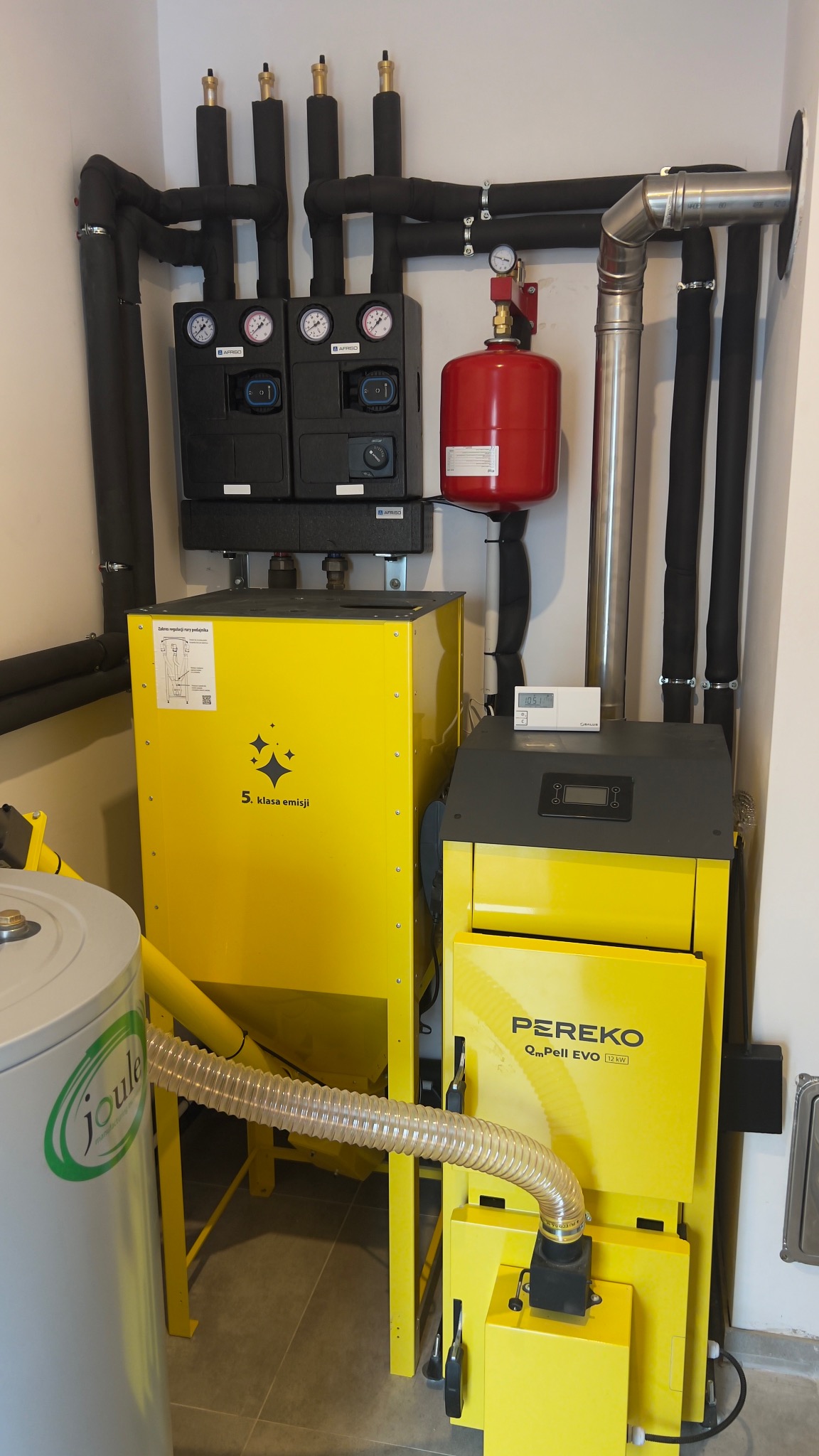 Żółty piec na pellet Pereko QmPell EVO 12 kW z czarnymi rurami i elementami instalacji grzewczej w kotłowni. Widoczny zbiornik Joule i czujnik temperatury.