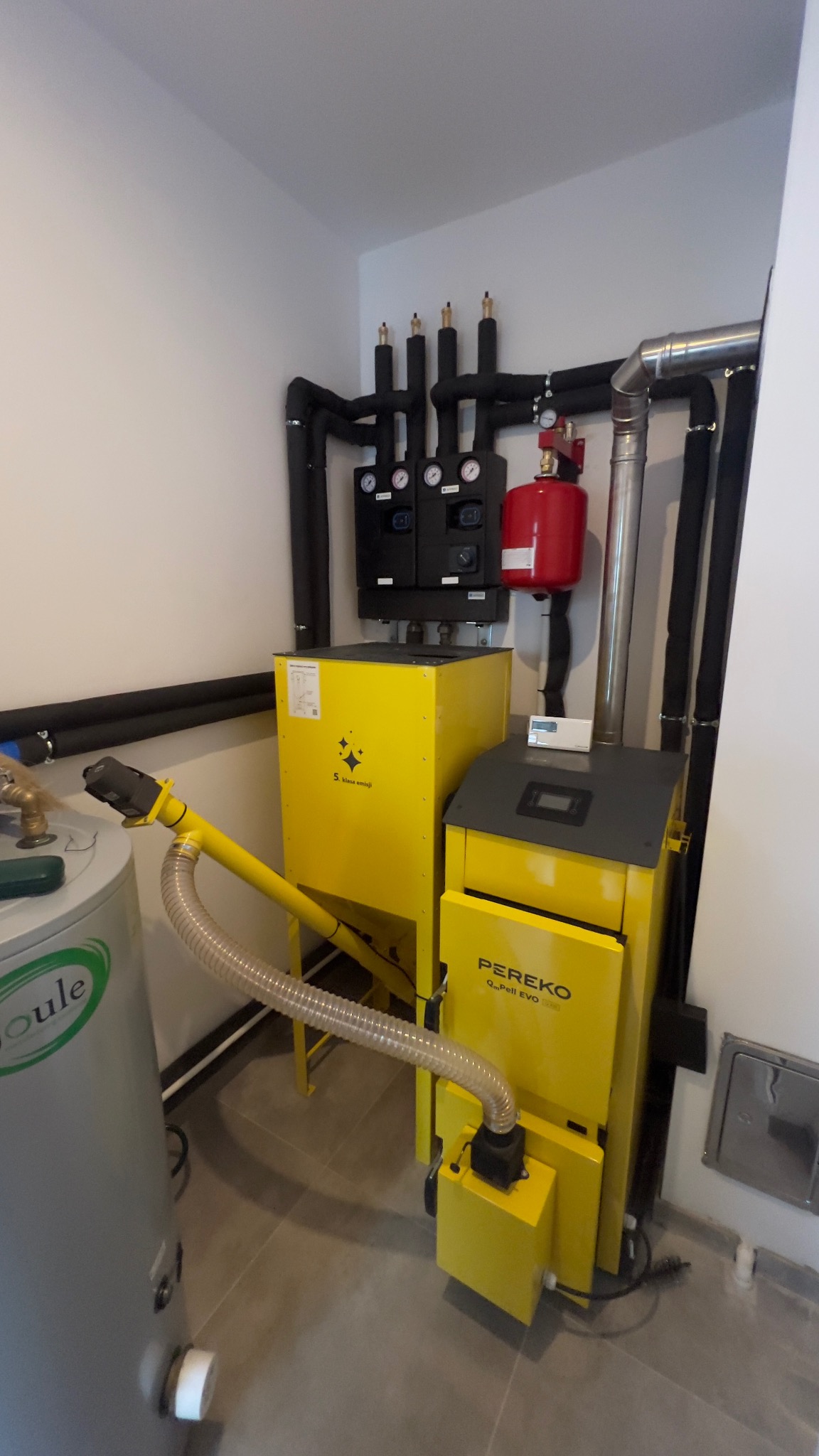 Żółty piec na pellet Pereko QmPeII EVO z podajnikiem, instalacja CO z czarnymi rurami w kotłowni. Widoczny zbiornik buforowy i osprzęt.