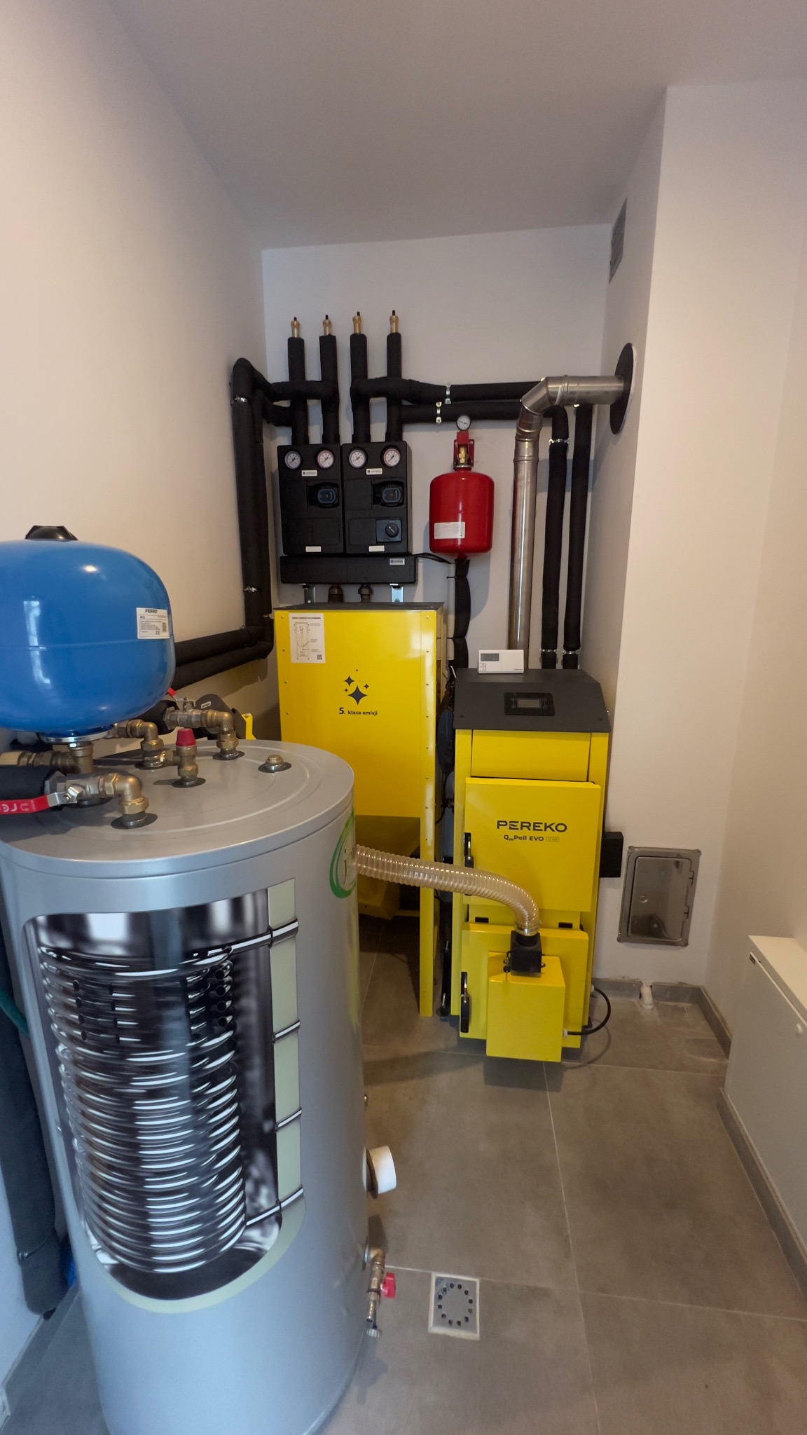 Żółty piec Pereko z instalacją CO, zbiornik z niebieskim naczyniem przeponowym, czarne rury i armatura w kotłowni. Widok na kompletną instalację grzewczą.