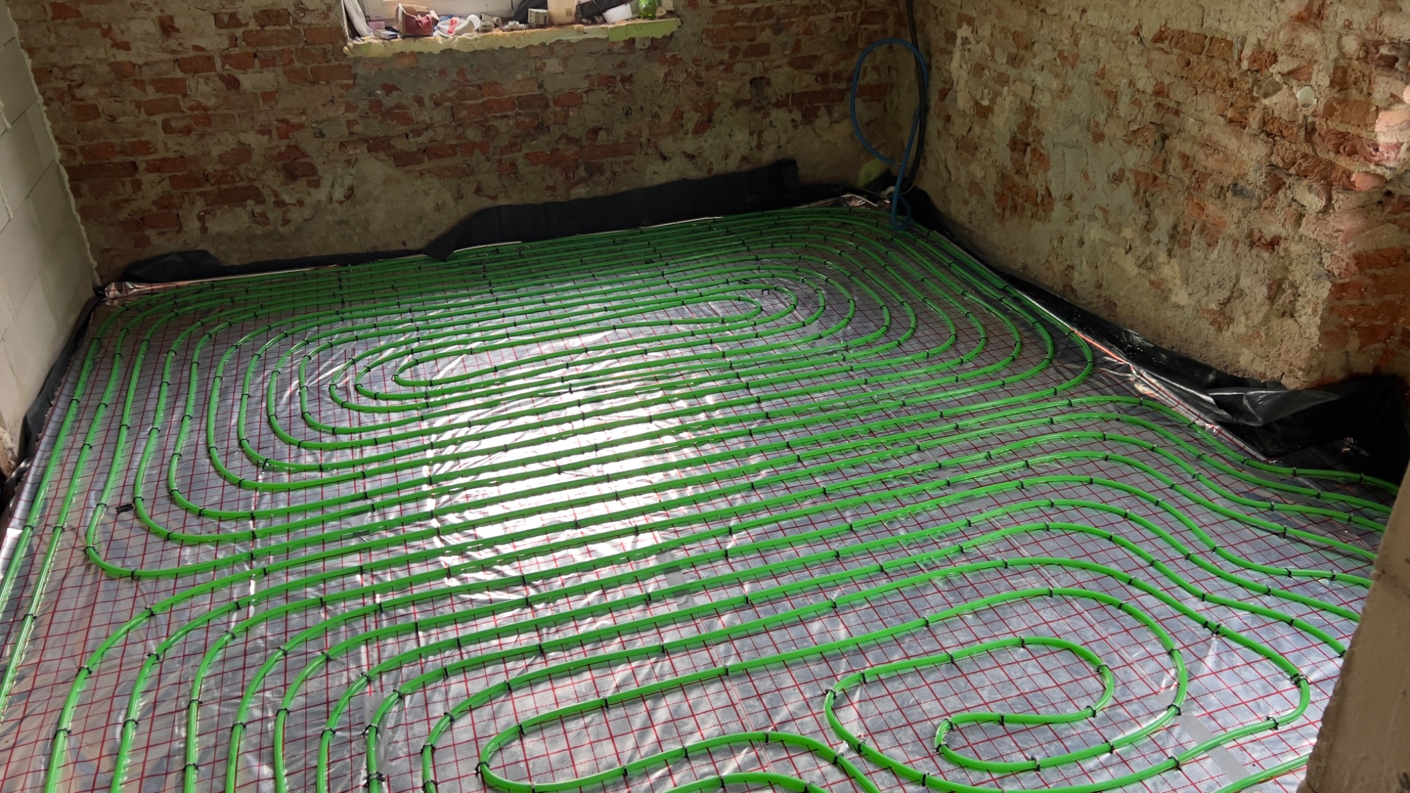 Zielone rury ogrzewania podłogowego ułożone na folii z czerwoną kratką, widoczne fragmenty surowej, ceglanej ściany. Instalacja w trakcie realizacji, przed wylaniem posadzki.