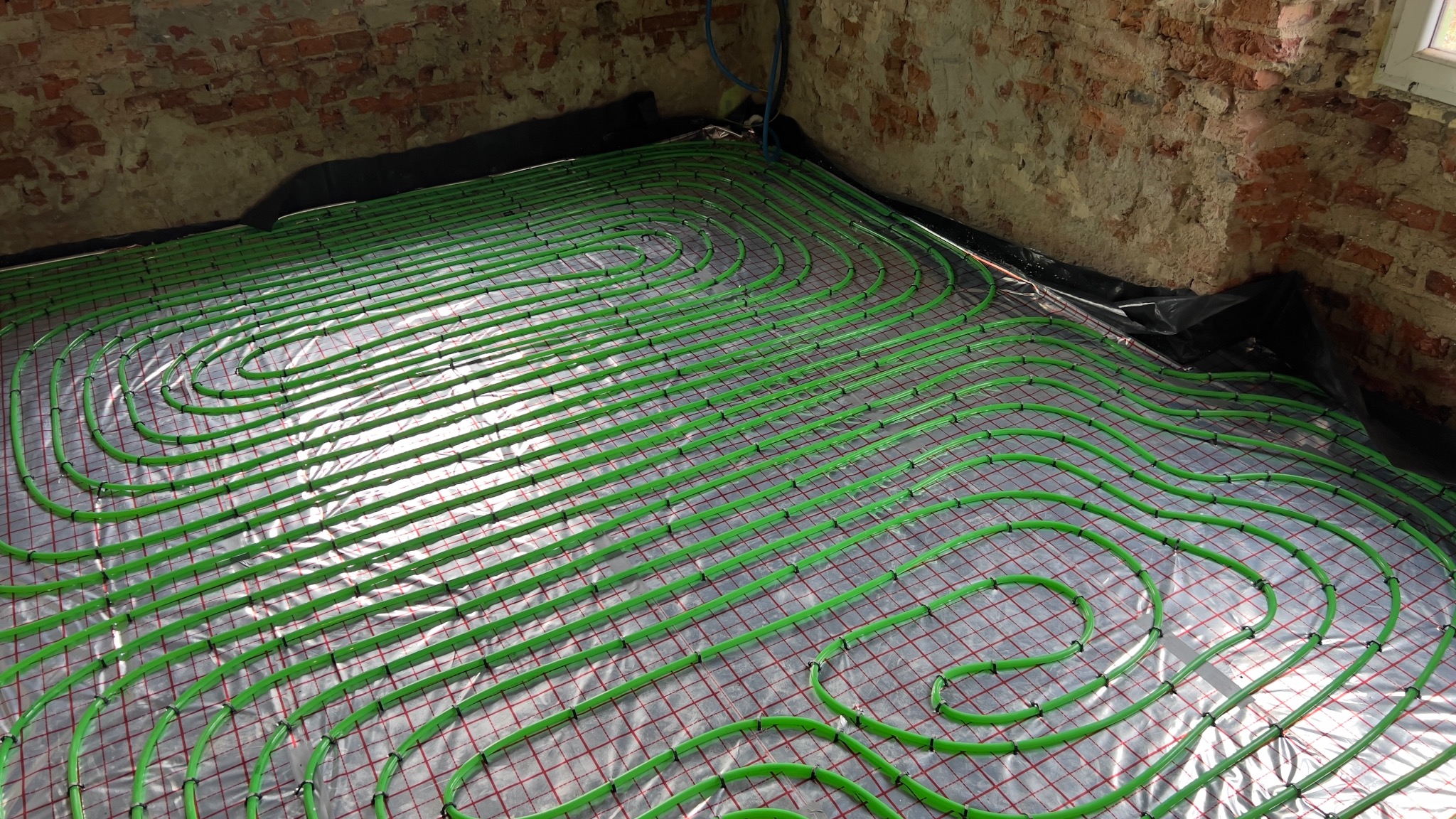 Zielone rury ogrzewania podłogowego ułożone spiralnie na folii z czerwoną siatką, widoczne fragmenty ceglanej ściany w surowym stanie. Instalacja przed zalaniem wylewką.