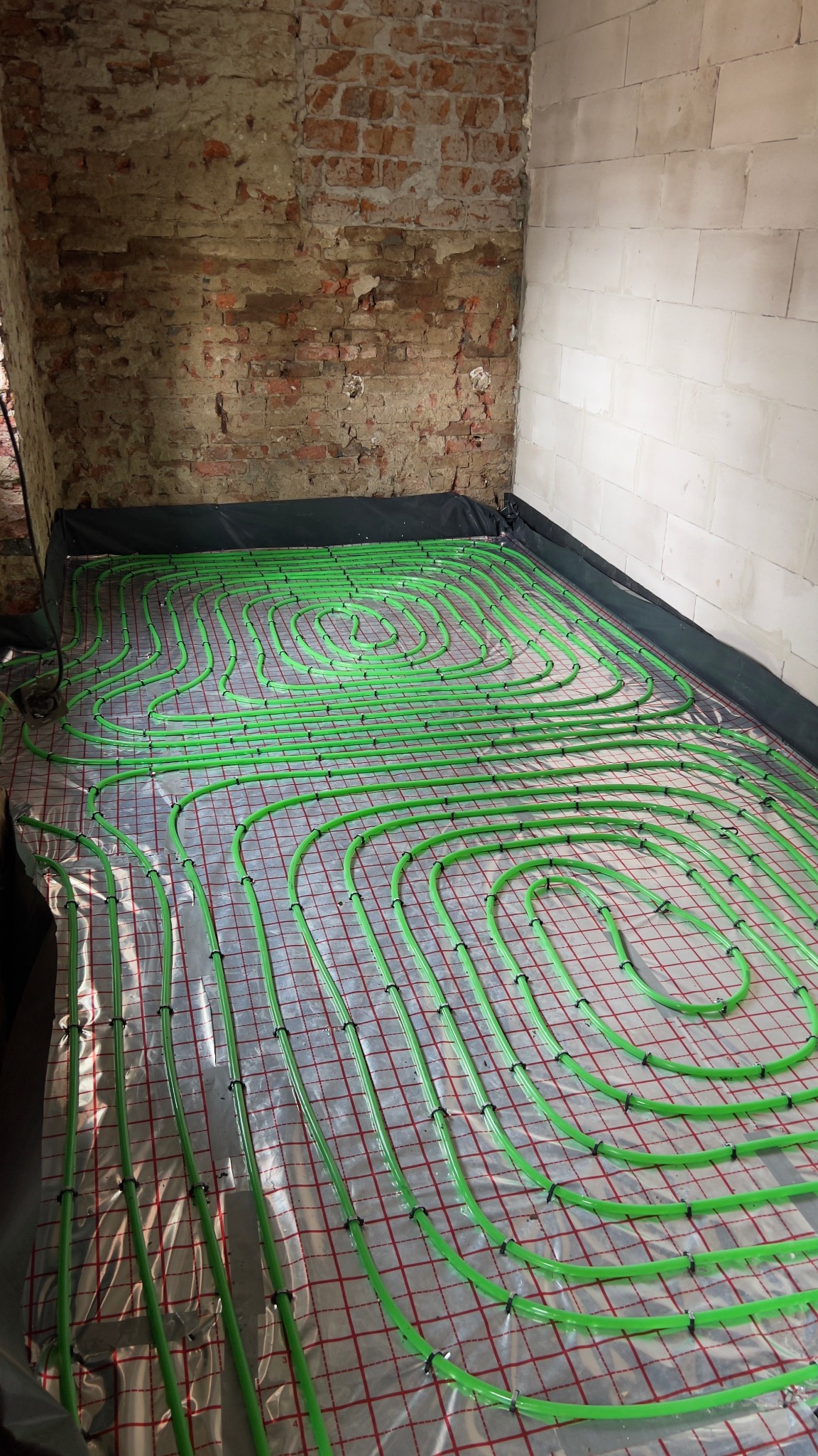 Zielone rury ogrzewania podłogowego ułożone na folii z siatką, widoczne fragmenty surowych ścian z cegły i bloczków. Instalacja przed wylaniem posadzki.
