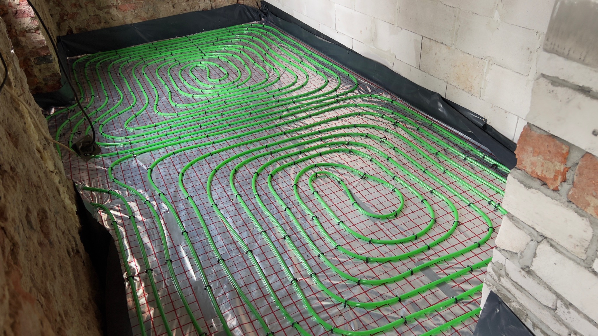 Zielone rury ogrzewania podłogowego ułożone spiralnie na folii z siatką, widoczne fragmenty ścian z cegły i bloczków. Instalacja w trakcie realizacji.