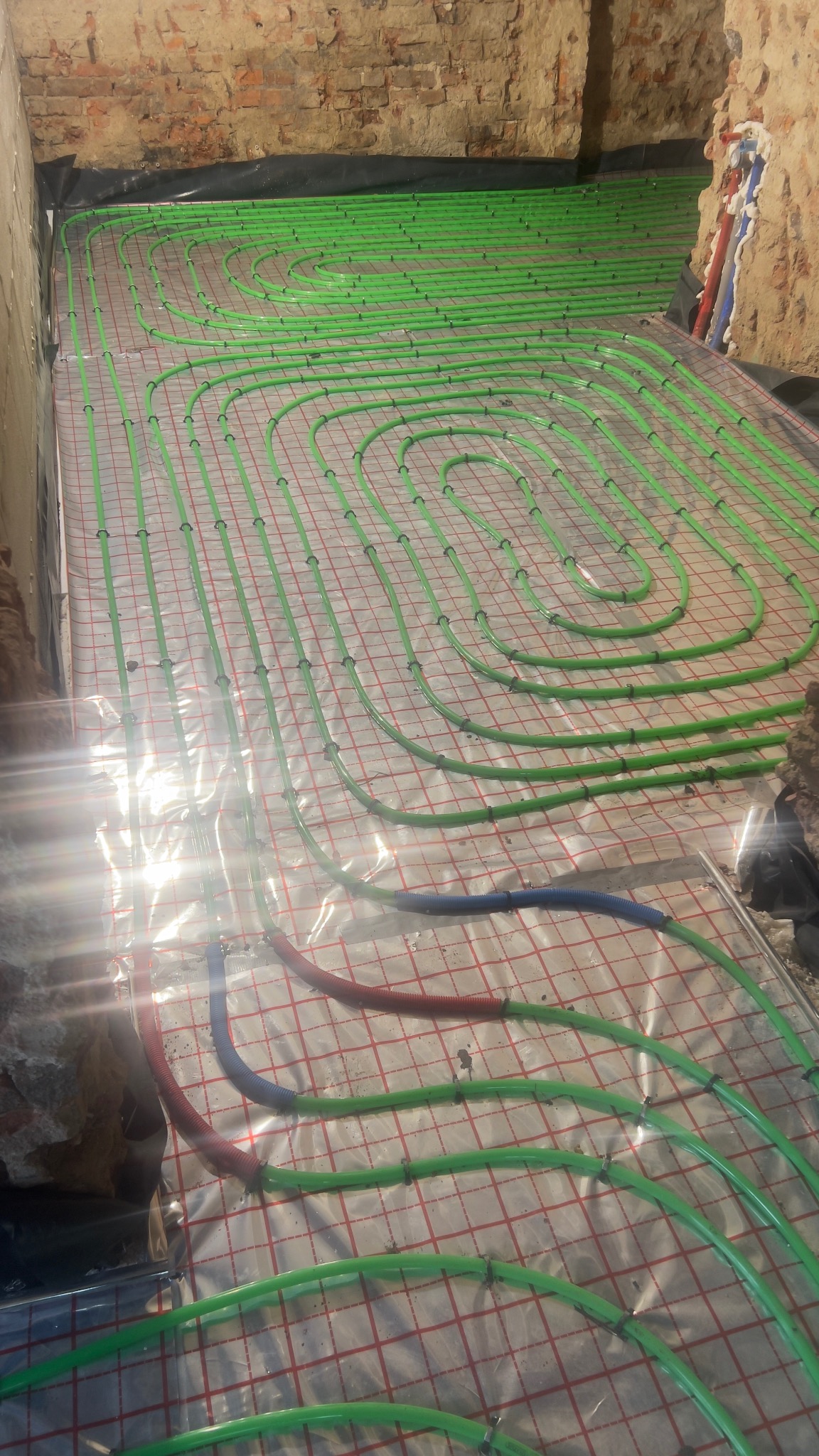 Zielone rury ogrzewania podłogowego ułożone spiralnie na folii z czerwoną siatką, widoczne fragmenty muru z cegły i inne rury w kolorze niebieskim i czerwonym.