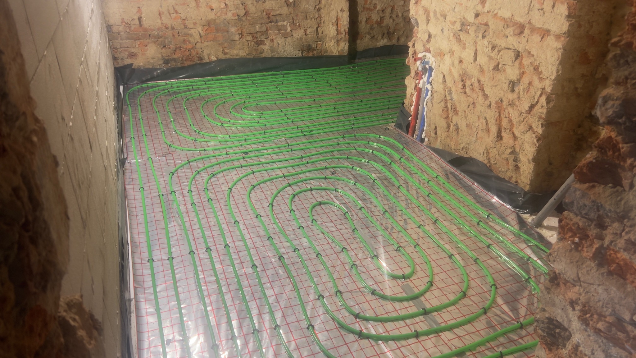 Ułożone, zielone rury ogrzewania podłogowego na folii, widoczne fragmenty ścian z cegły i zaprawy. Instalacja w trakcie realizacji, przed wylaniem posadzki.