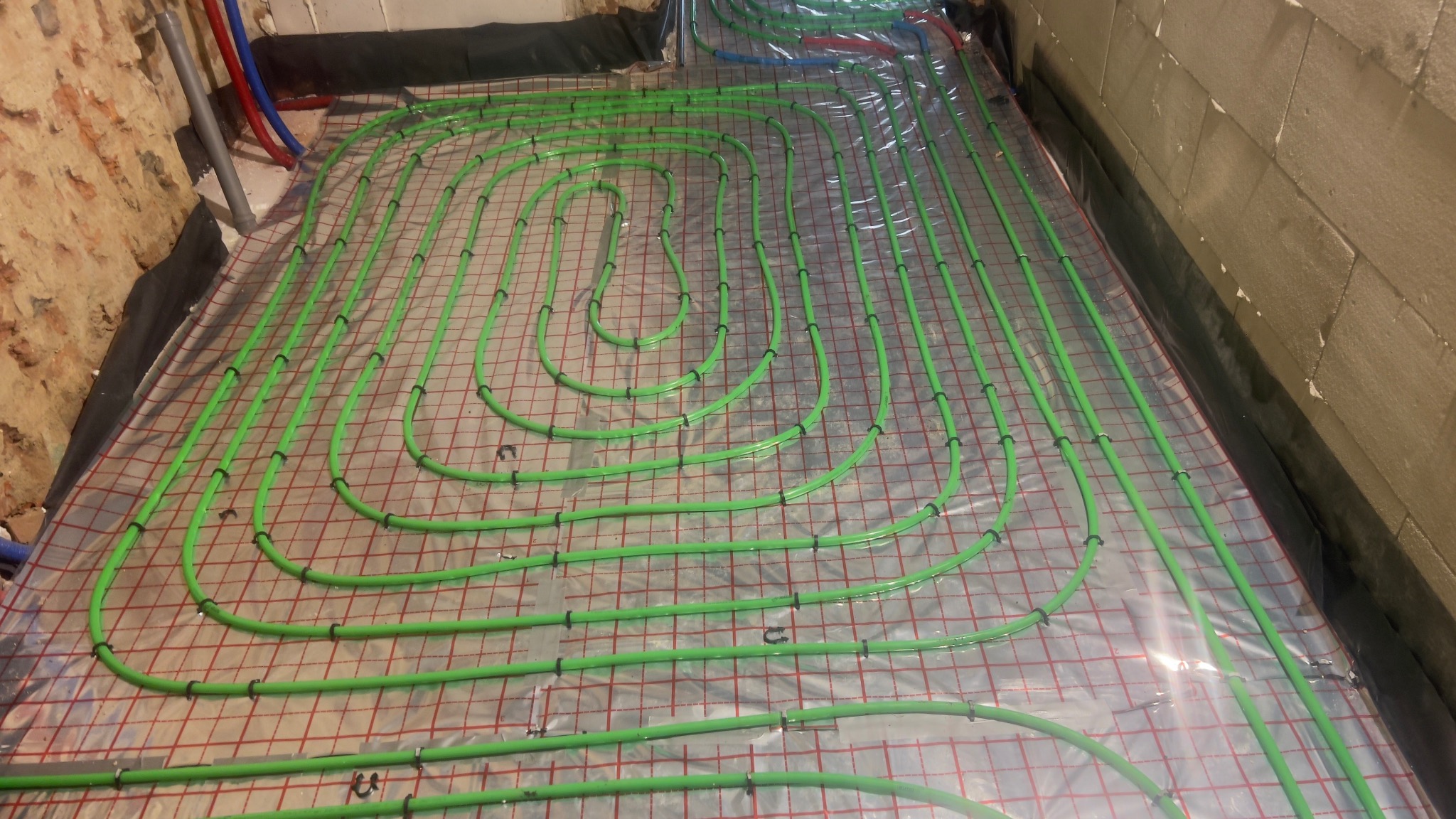 Zielone rury ogrzewania podłogowego ułożone na folii z czerwoną siatką, przymocowane czarnymi uchwytami. Widoczne fragmenty ścian i rur instalacji.