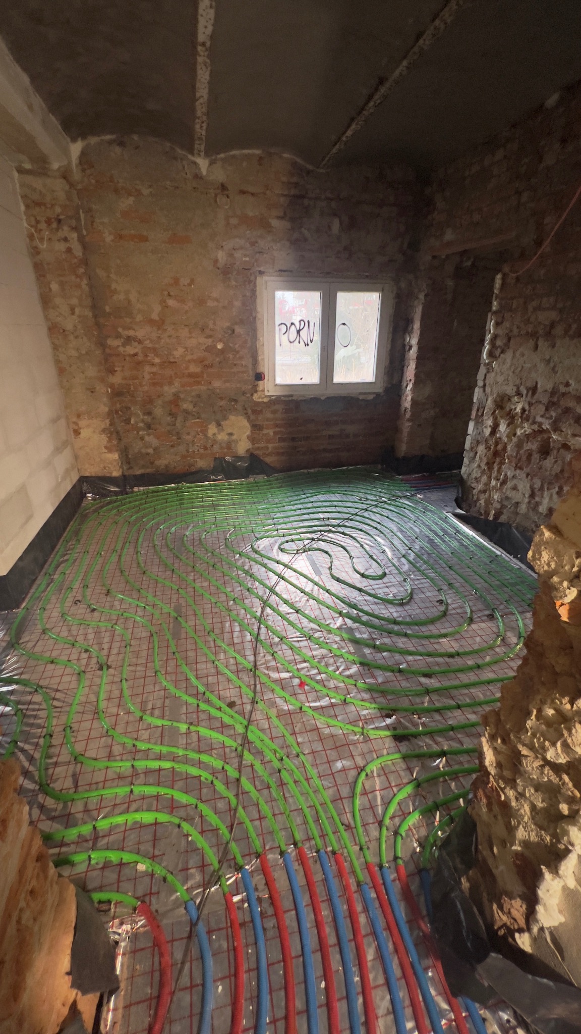 Instalacja ogrzewania podłogowego z rurami w kolorze zielonym, czerwonym i niebieskim, ułożonymi na folii z siatką, w surowym wnętrzu z ceglanymi ścianami i oknem.