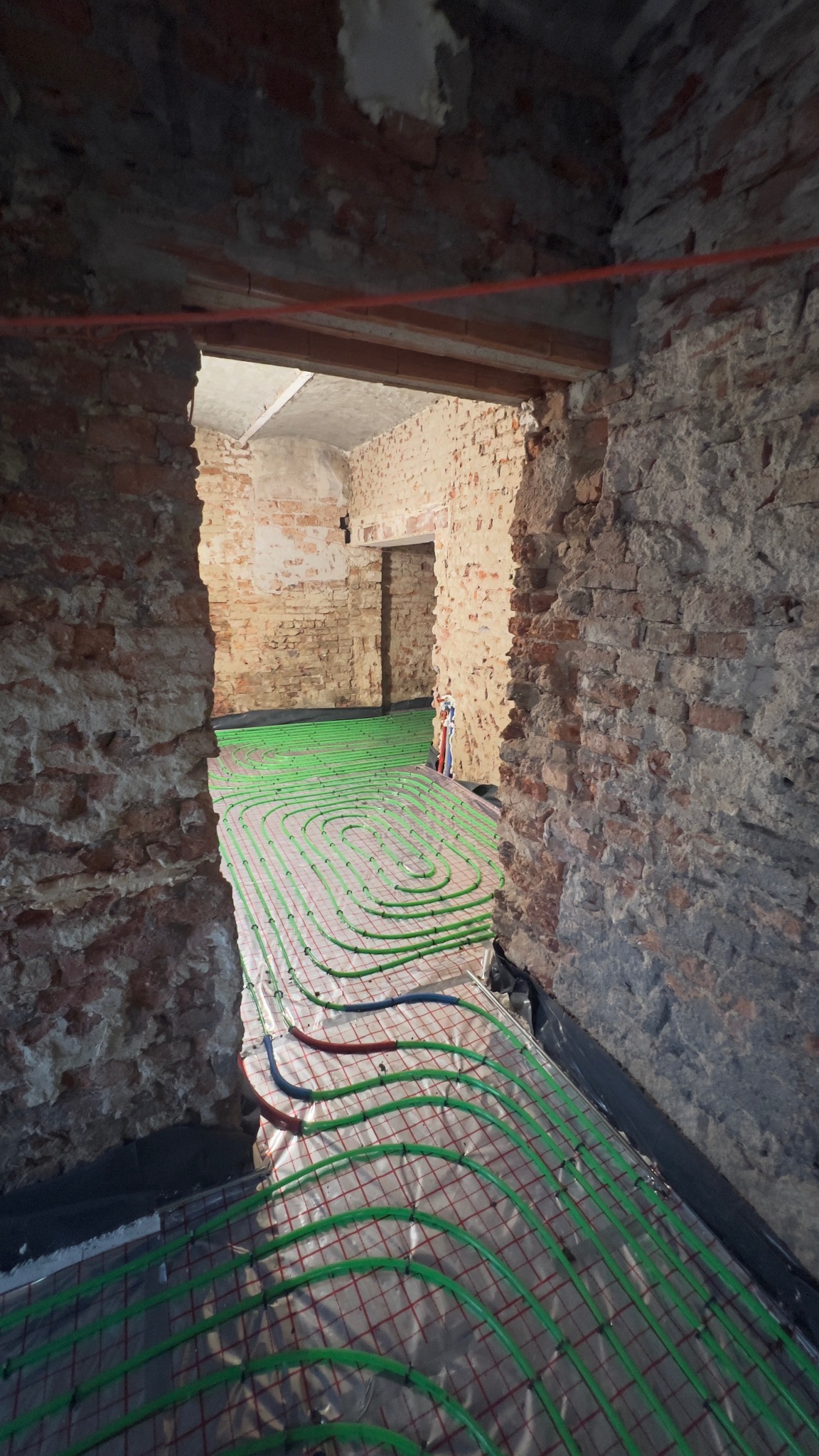 Zielone rury ogrzewania podłogowego ułożone w surowym wnętrzu z ceglanymi ścianami. Widok przez otwór drzwiowy, instalacja na folii z czerwoną siatką.