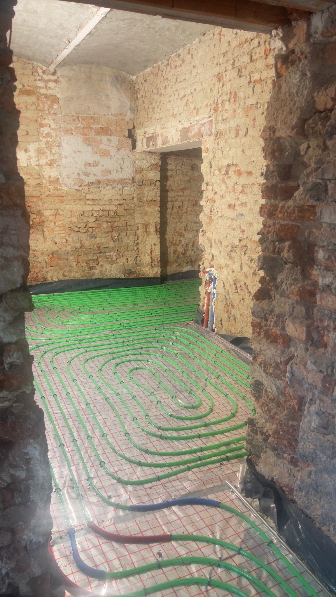 Zielone rury ogrzewania podłogowego ułożone na folii w surowym wnętrzu z ceglanymi ścianami. Widok przez otwór w ścianie, ukazujący etap instalacji.