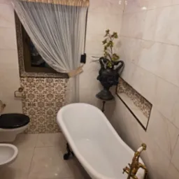 Elegancka łazienka w stylu retro z wolnostojącą wanną na lwich łapach, zdobioną armaturą i marmurkowymi płytkami. Okno z ozdobną zasłoną.