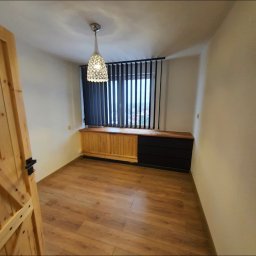 N-techniczne - Jasne panele podłogowe, drewniana komoda pod oknem z żaluzjami w pokoju. Widoczna drewniana rama drzwi z klamką. Lampa sufitowa w stylu nowoczesnym.