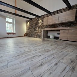 N-techniczne - Jasne panele podłogowe z efektem drewna w przestronnym wnętrzu z ceglaną ścianą, drewnianymi szafkami i oknem witrażowym. Widoczne czarne belki stropowe.