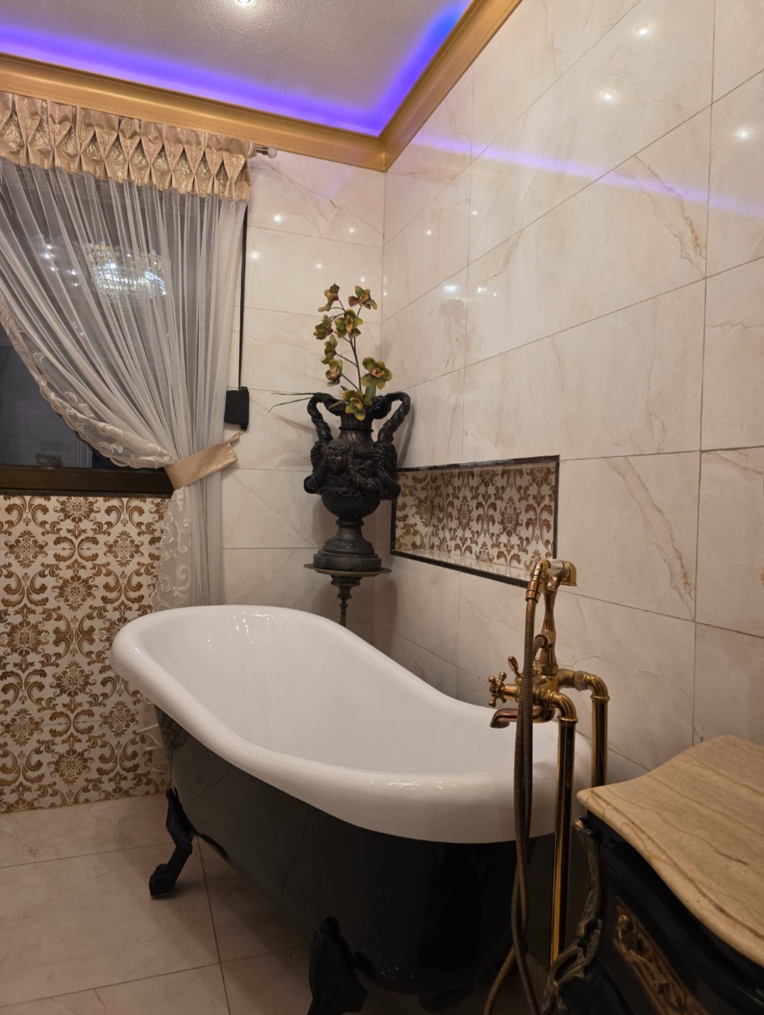 Elegancka łazienka z wolnostojącą wanną na lwich nóżkach i złotą armaturą. Marmurowe płytki, dekoracyjna waza z kwiatami i podświetlany sufit tworzą luksusowy klimat.